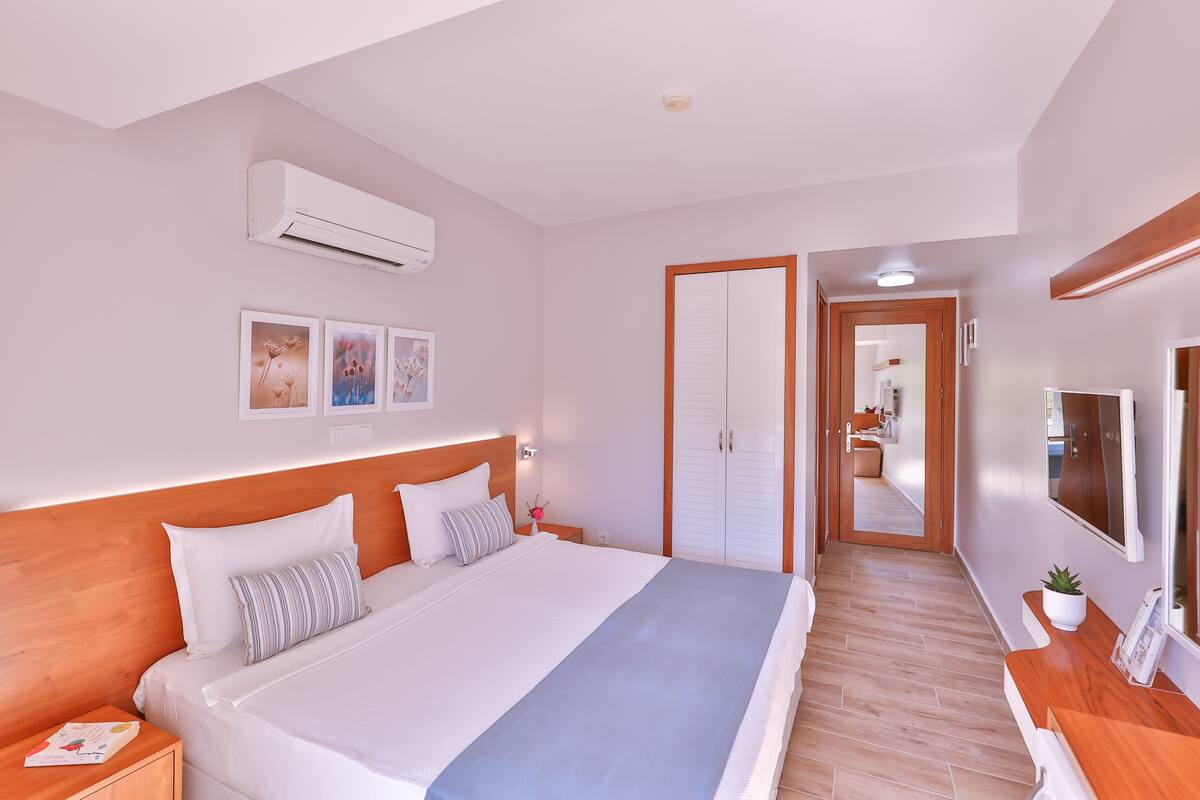 A modern hotel room with a large bed, wooden headboard, white walls, and a hallway leading to a bathroom.