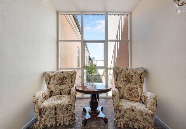 Une pièce d’angle avec deux fauteuils à motifs floraux, une table ronde en bois et une grande fenêtre donnant sur des immeubles.
