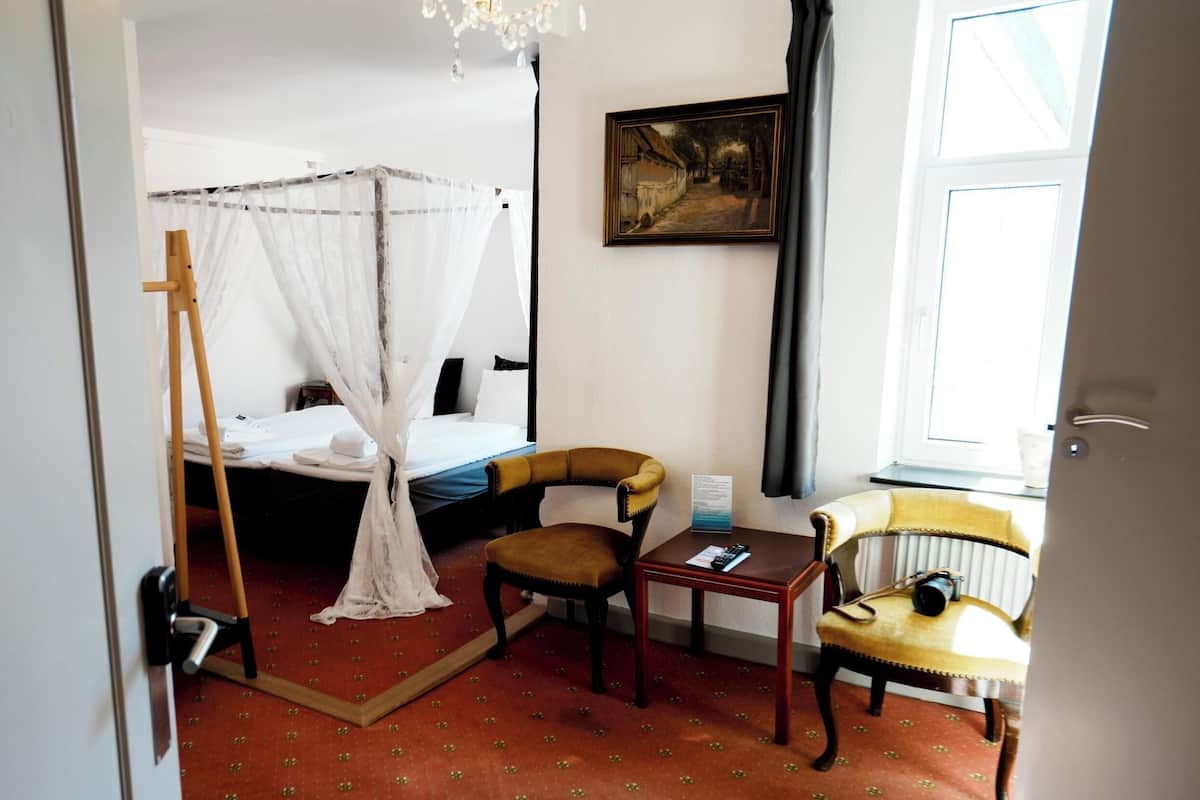 A room with a bed, a chair, a small table, a radiator, and a painting on the wall.