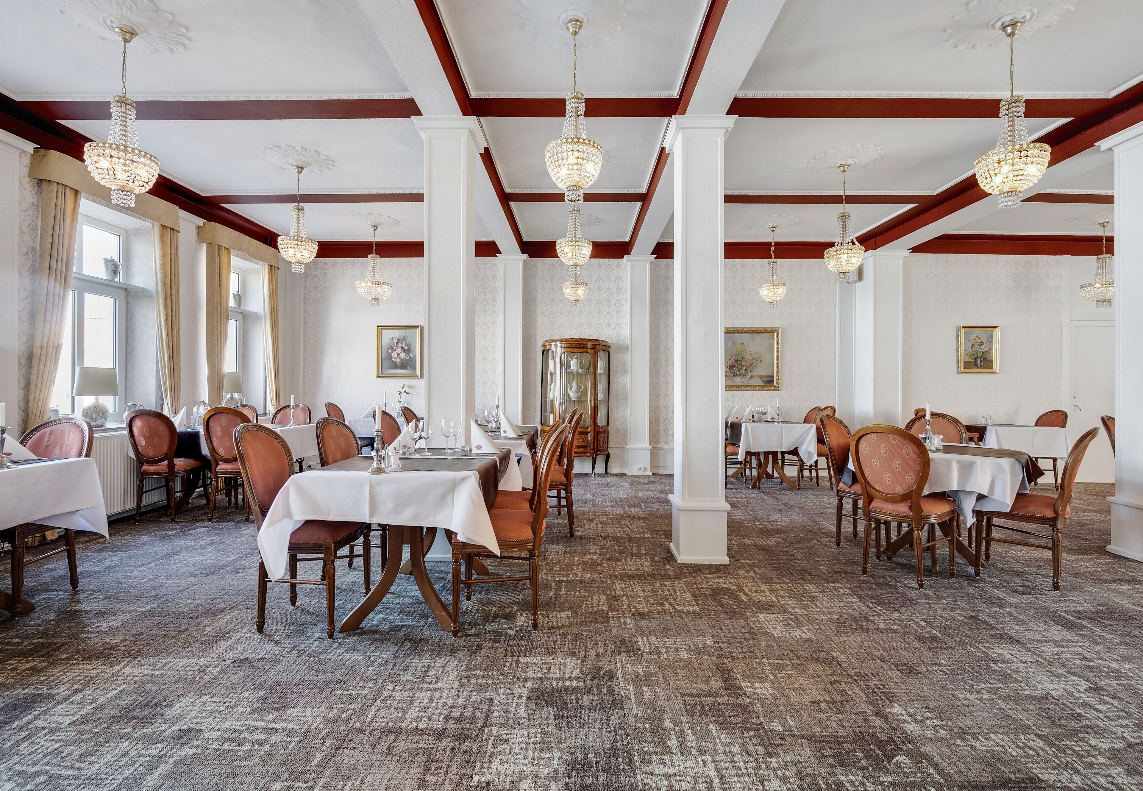 A dining area with tables set for service, wooden chairs, chandeliers, and a carpeted floor.