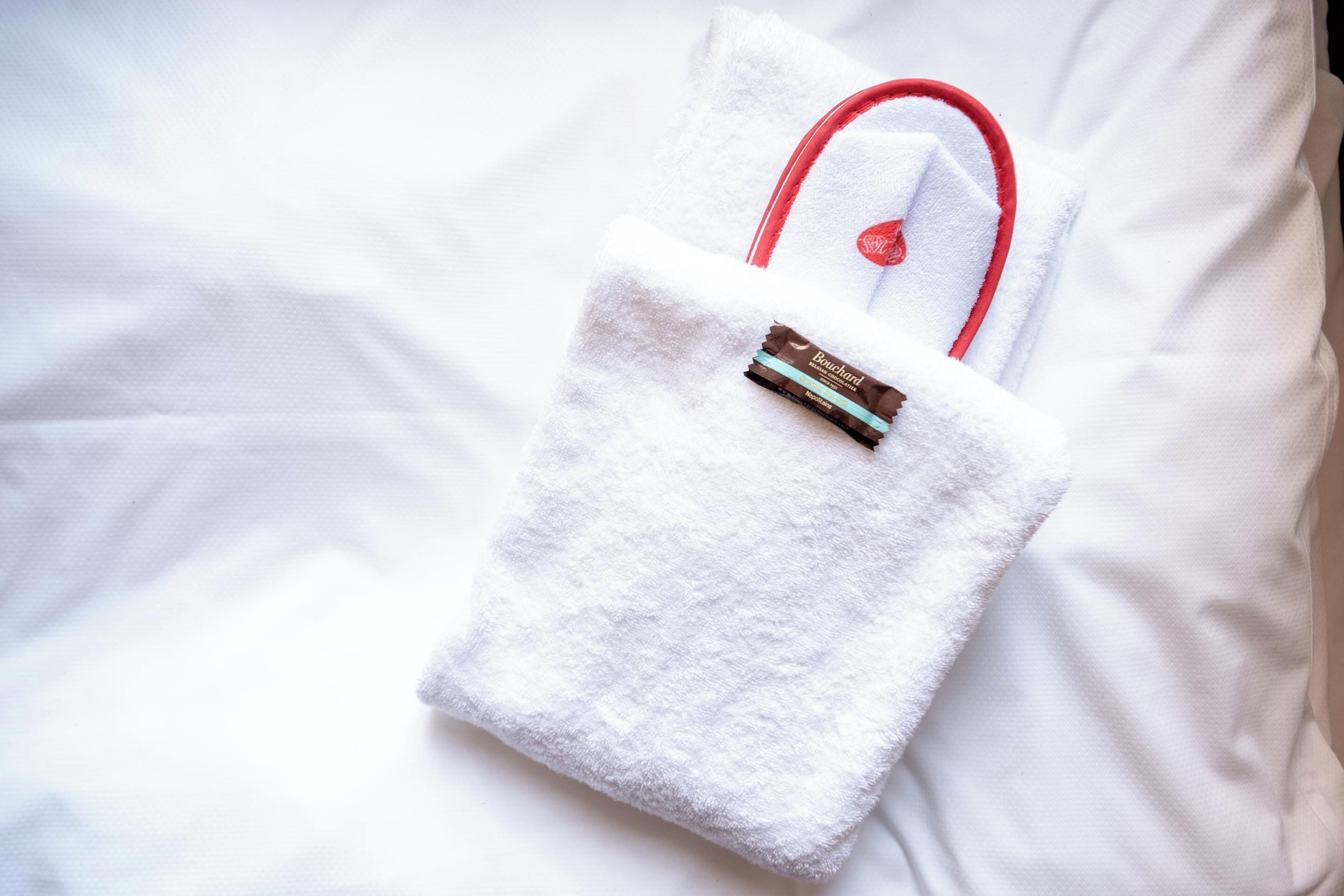 Una toalla blanca con asa roja y una barra de chocolate sobre una cama blanca.