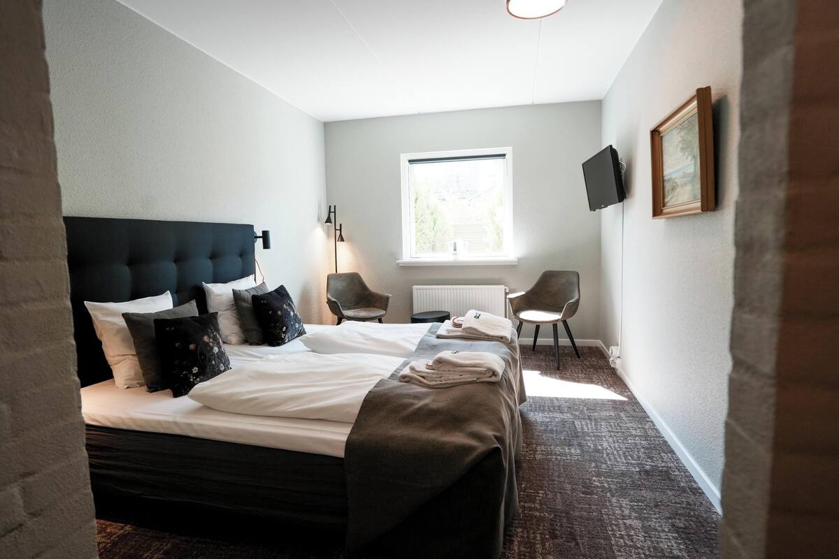 A hotel room with a large bed, two chairs, a TV, and a window.
