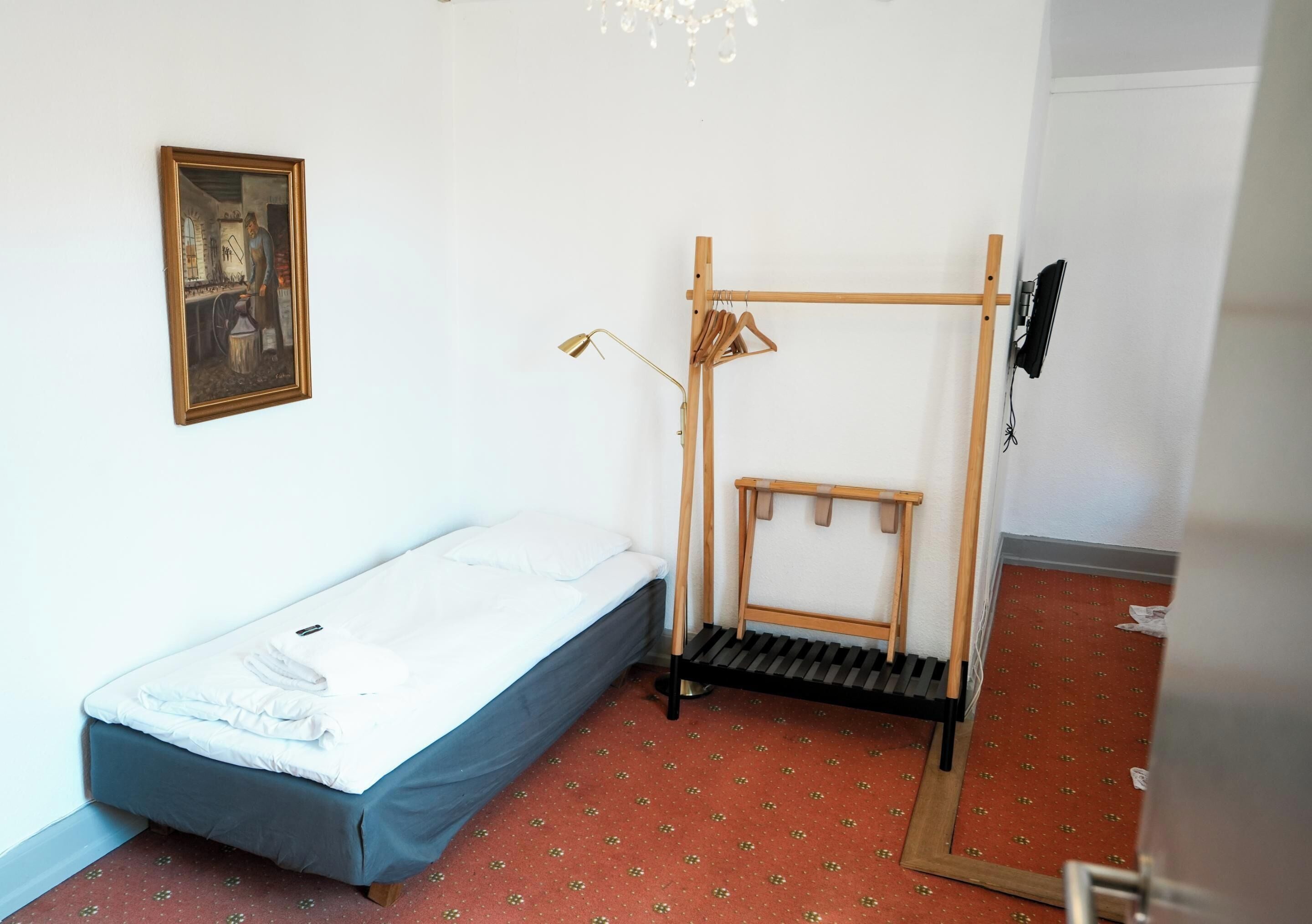 A small, simple room with a single bed, a wooden rack, and a framed picture on the wall.