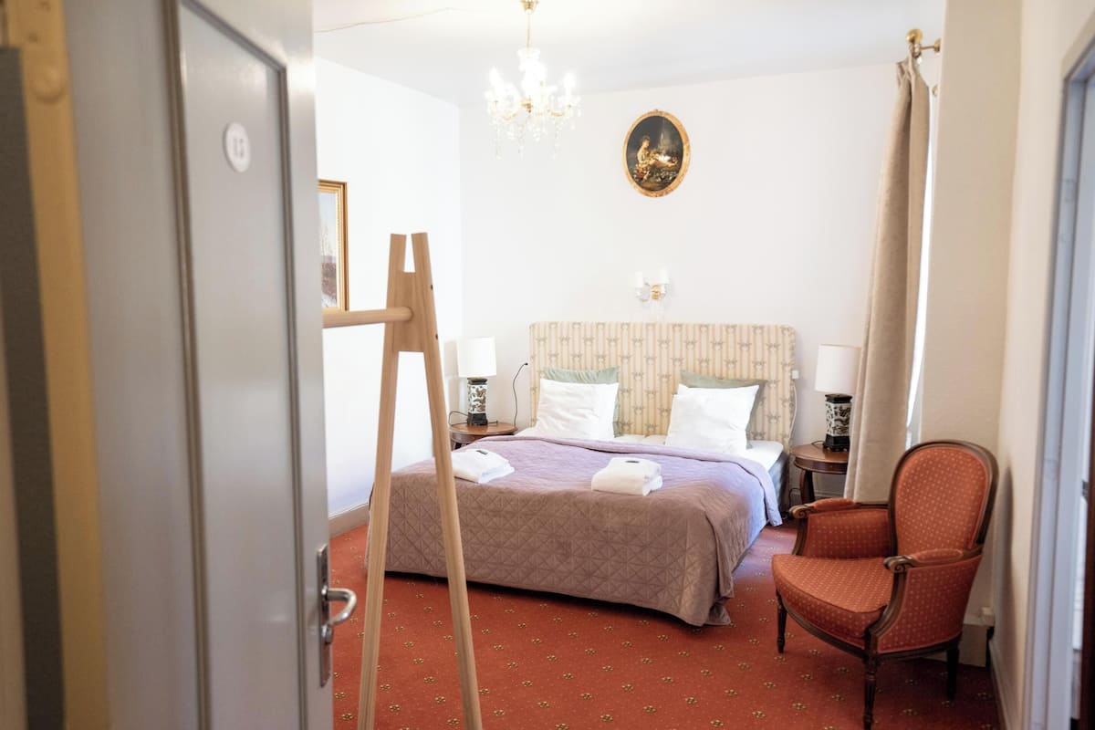 A hotel room with a bed, a chair, a desk, a chandelier, and a painting on the wall.