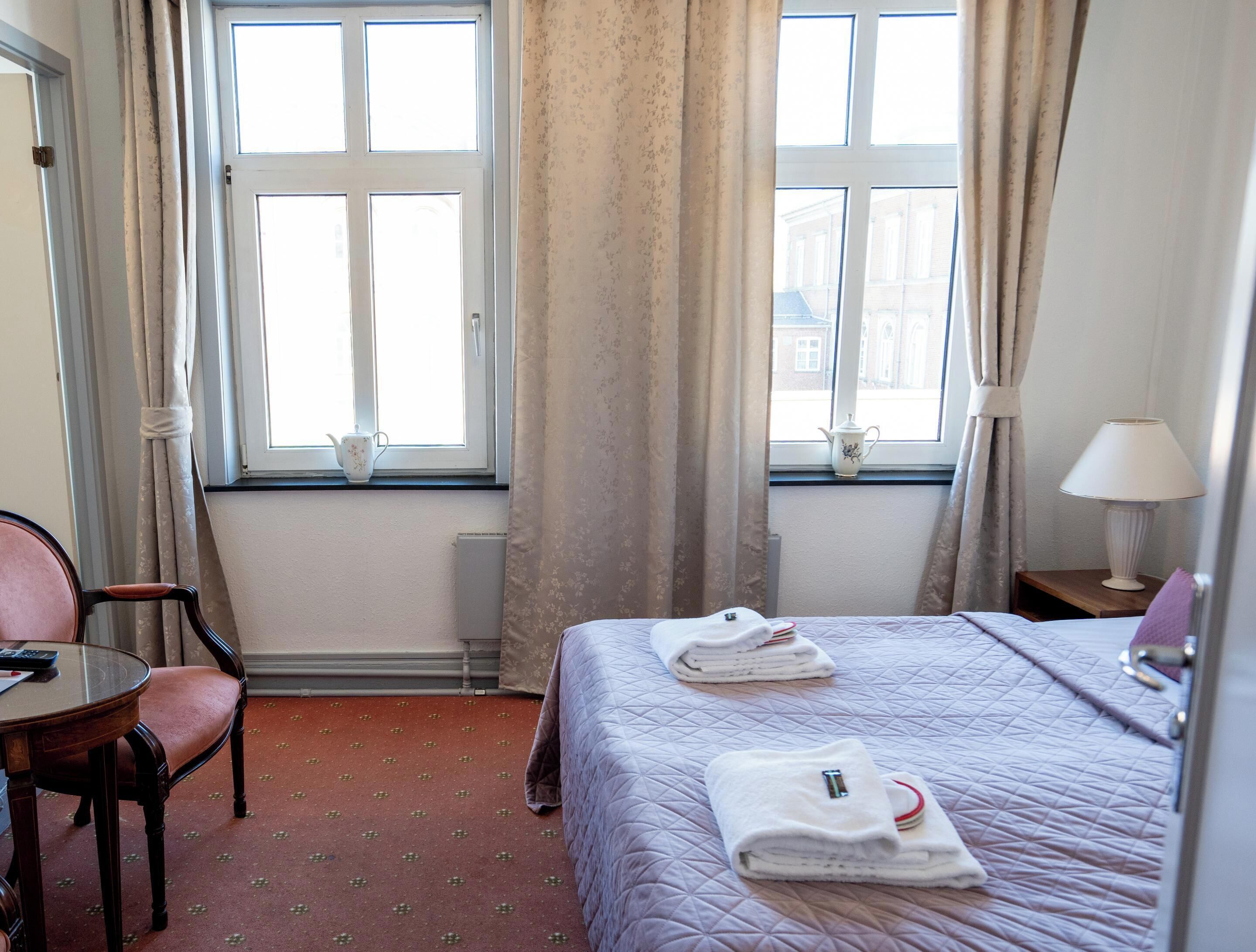 Quarto de hotel com cama, poltrona, mesa, abajur e duas janelas com cortinas.