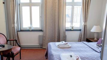 Quarto de hotel com cama, poltrona, mesa, abajur e duas janelas com cortinas.