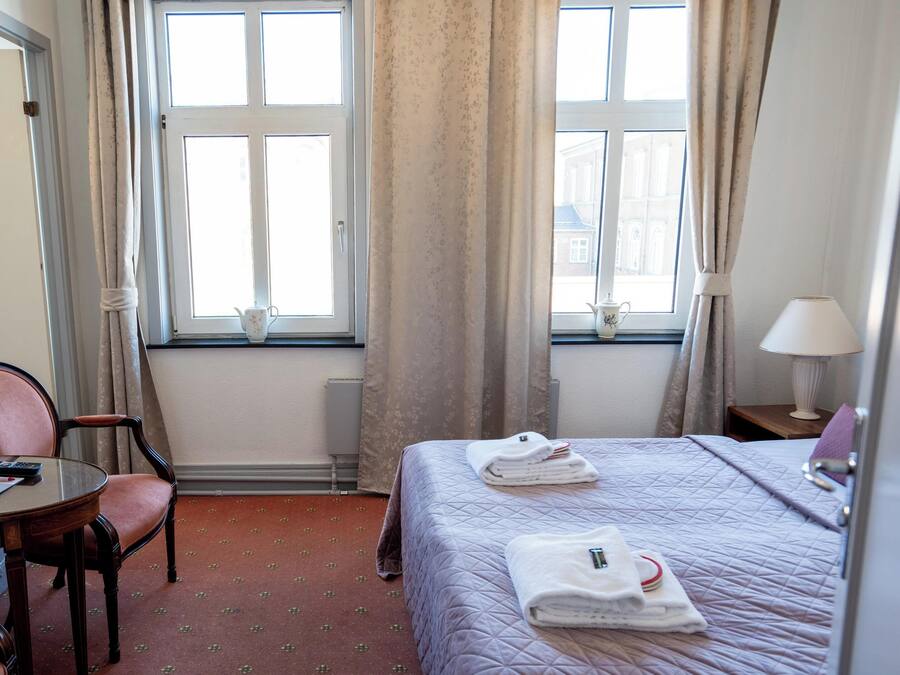 Quarto de hotel com cama, poltrona, mesa, abajur e duas janelas com cortinas.