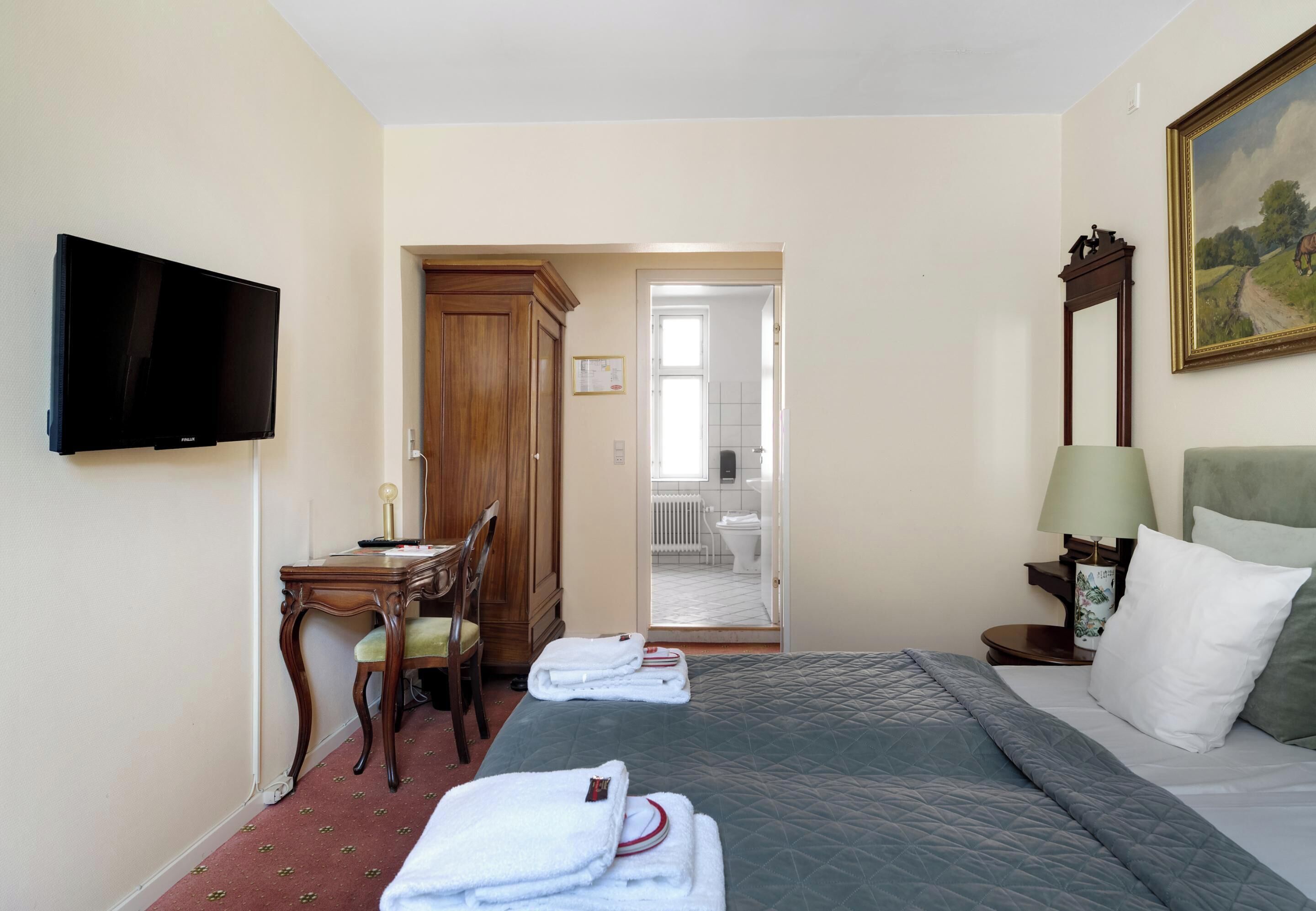 A hotel room with a bed, a desk, a chair, a television, and a painting on the wall.