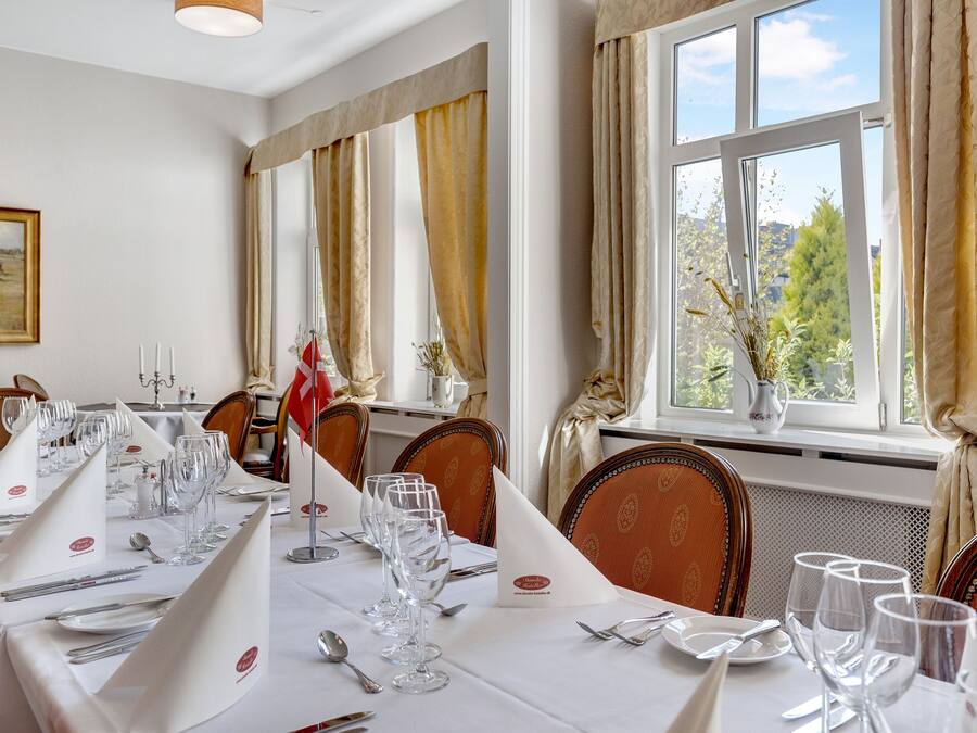 Une salle à manger avec une longue table dressée pour un repas, agrémentée de nappes blanches, de couverts disposés avec soin et de verres à vin.