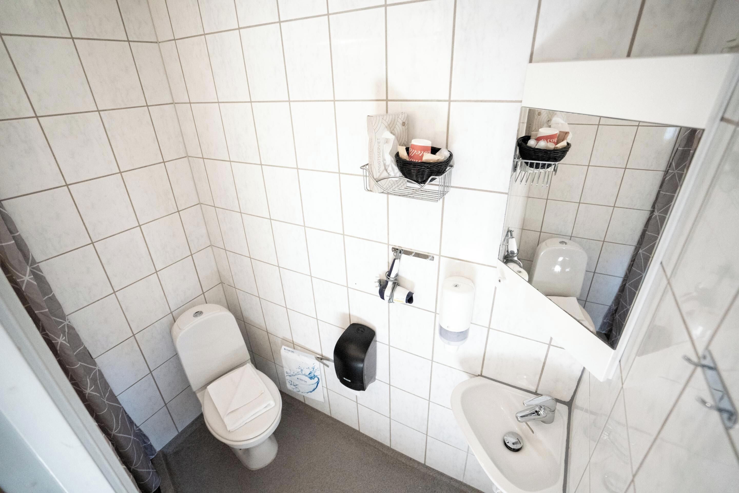 A small, tiled bathroom with a toilet, sink, and a mirror. There are shelves with toiletries and a paper towel dispenser.