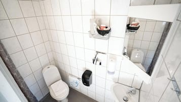 A small, tiled bathroom with a toilet, sink, and a mirror. There are shelves with toiletries and a paper towel dispenser.