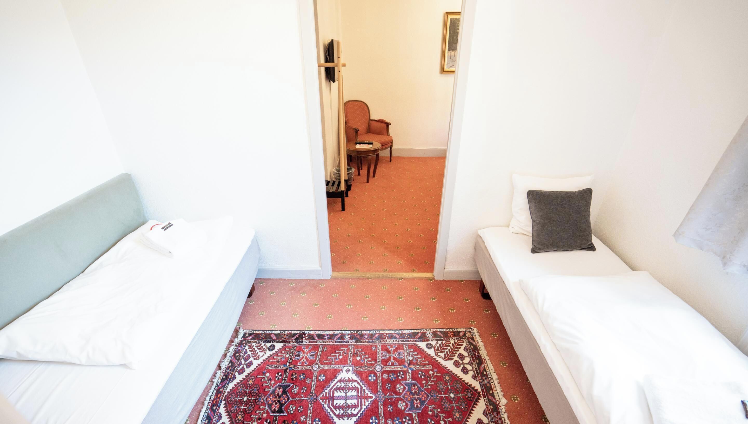 A small, well-lit room with two single beds, a red patterned rug, and a plain white wall.