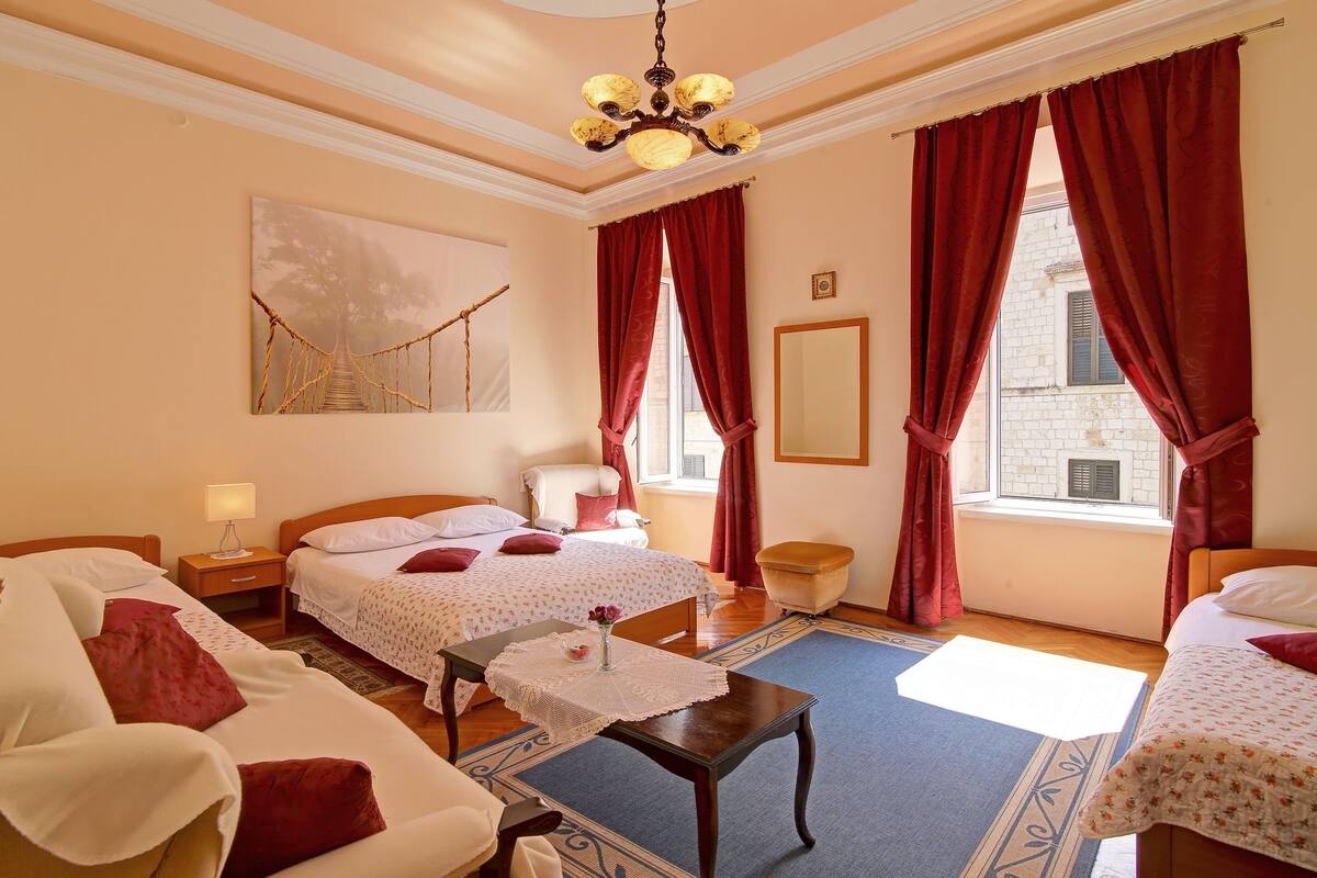 Habitación de hotel con dos camas, una mesita de noche de madera, una mesa con mantel de encaje, un candelabro y un ventanal grande con cortinas rojas.