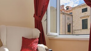 Una habitación con un sofá blanco, un cojín rojo, un tapete estampado y vistas a edificios a través de una ventana con cortinas rojas.
