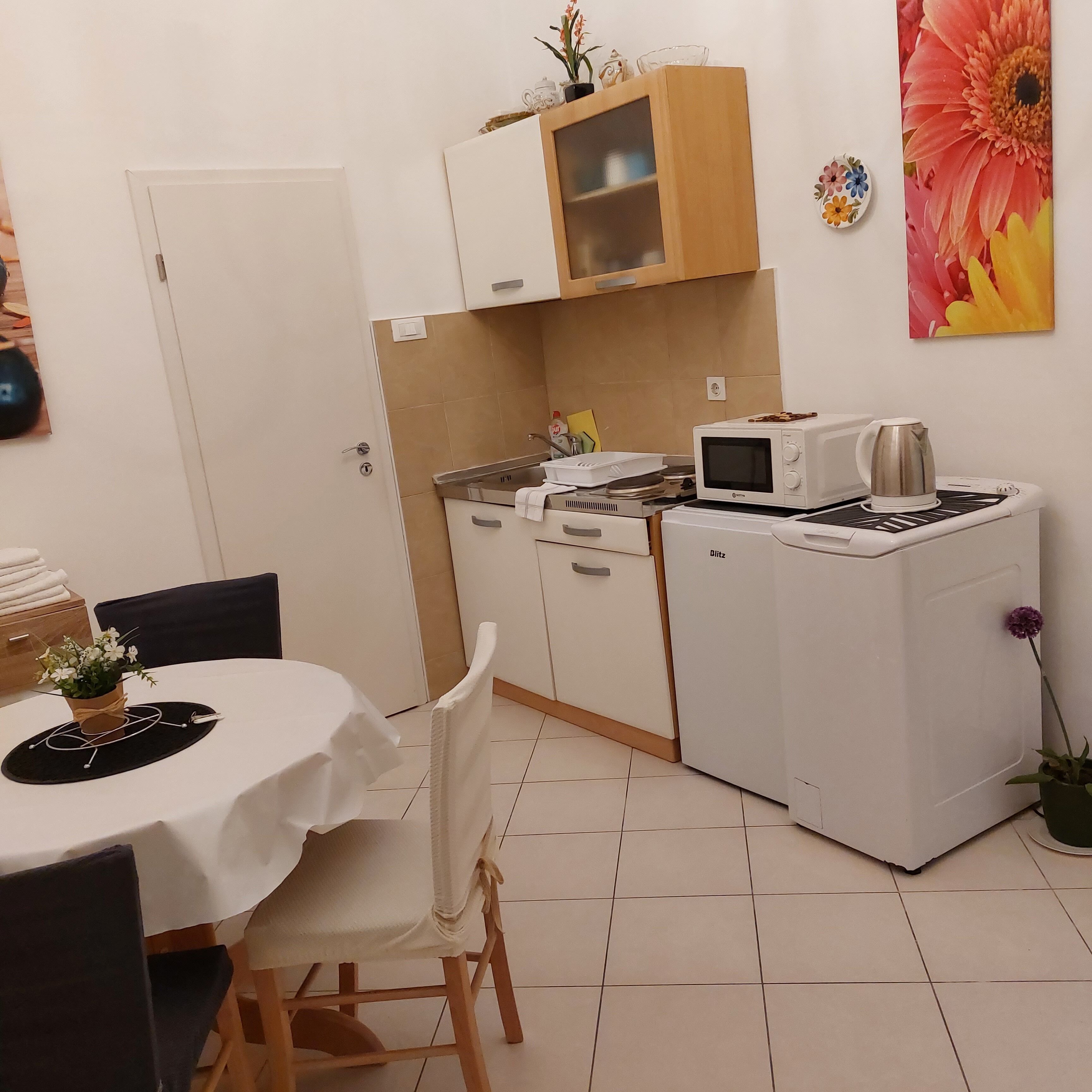 Una cocina compacta con microondas, hervidor y un refrigerador pequeño. Cuenta con una mesa de comedor con sillas y una planta en maceta.