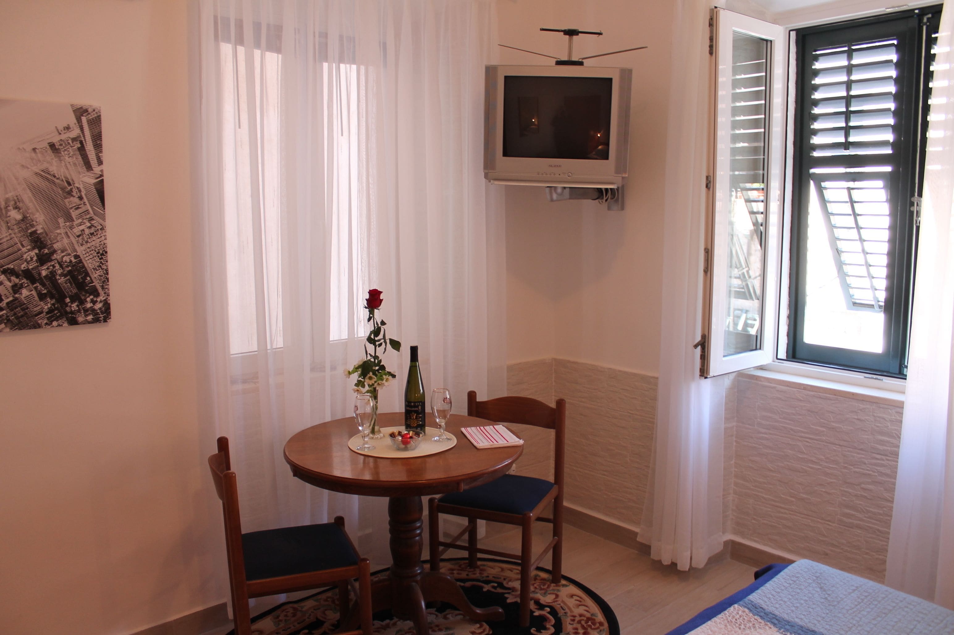 A dining area with a round table, two chairs, a television mounted on the wall, and a window with blinds.