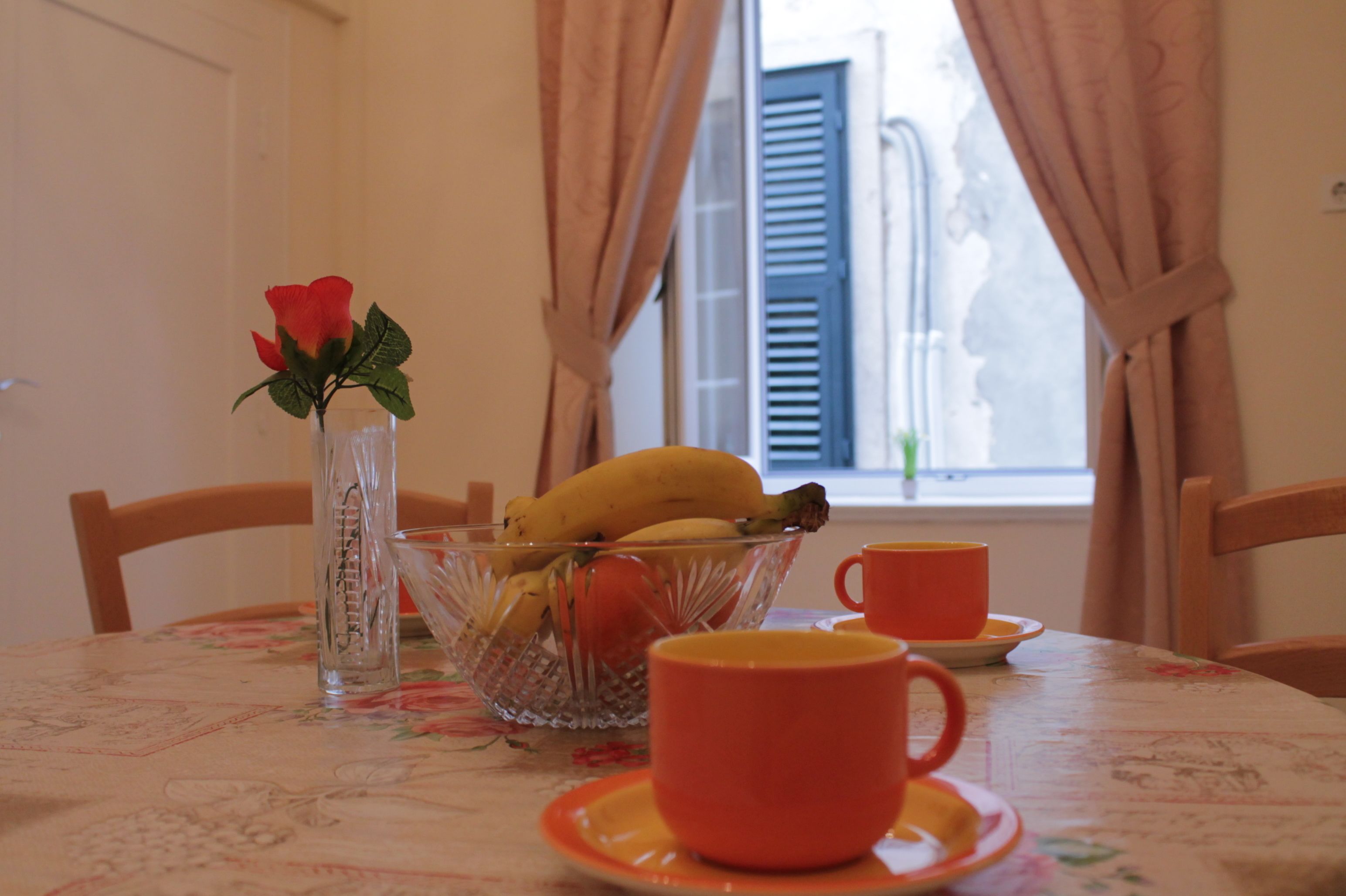 A dining table with a floral tablecloth, a glass bowl with bananas and a red rose, and a red cup and saucer.