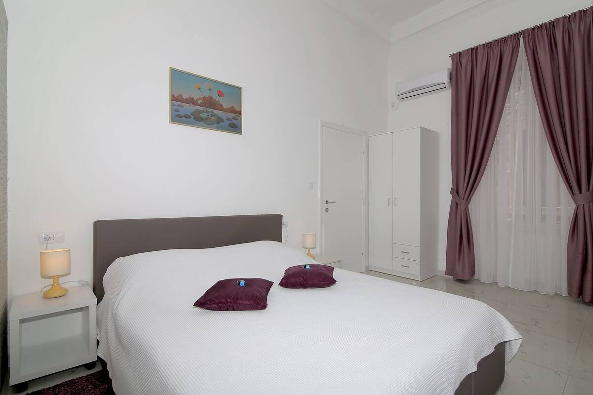 Una cama bien hecha con dos almohadas, una lámpara de noche, un cuadro en la pared y un armario.