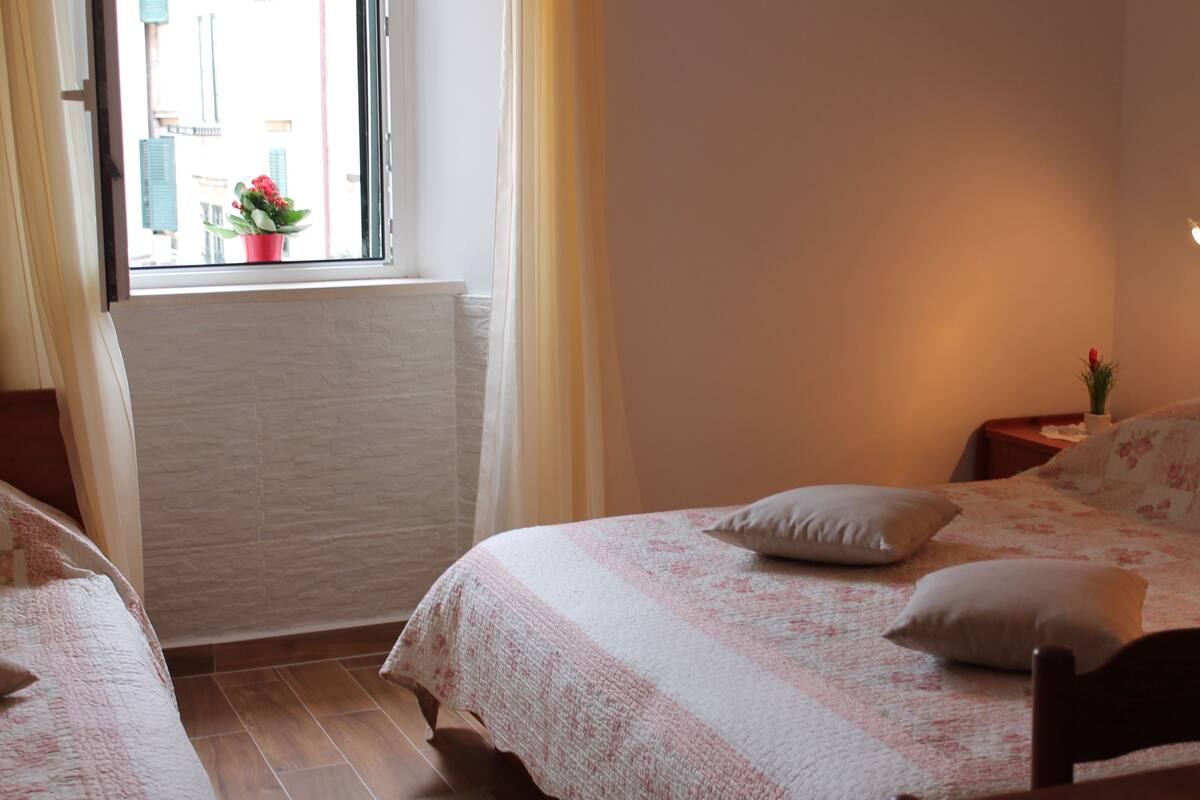 Un dormitorio con dos camas, una ventana que ofrece vistas a edificios, un radiador y una mesita de noche con lámpara.