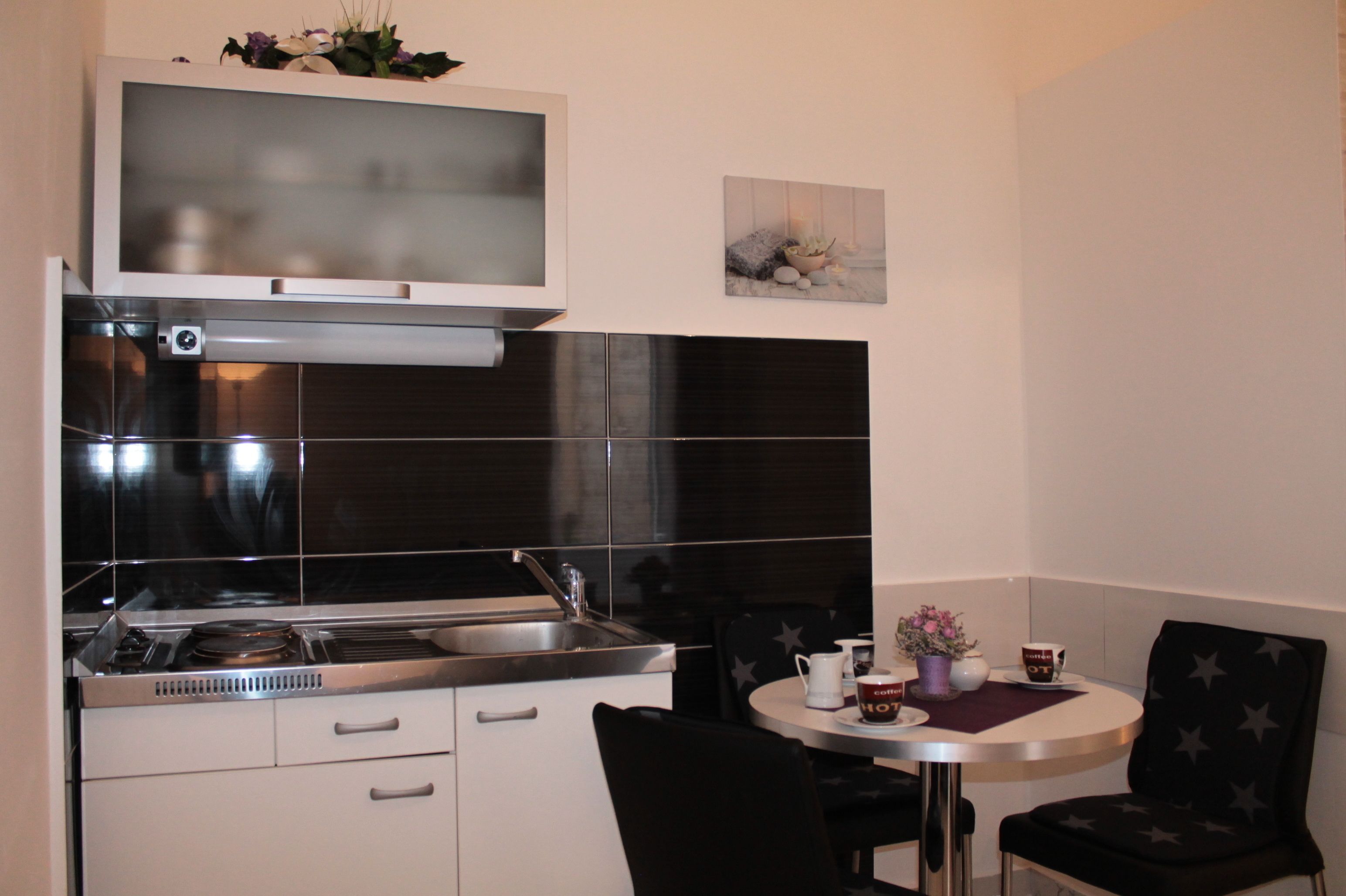 A compact kitchen with a stove, sink, and white cabinets. There is a small dining table with chairs and a decorative item on the wall.