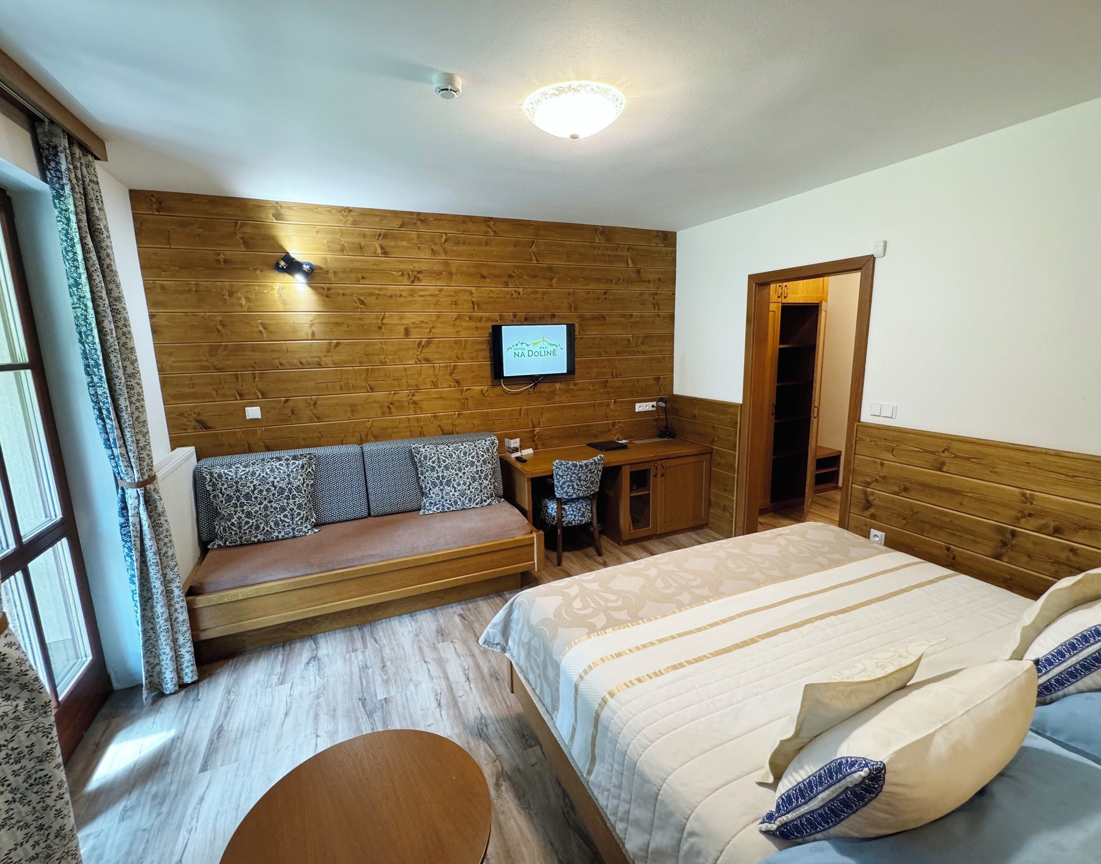 A hotel room with a wooden wall, a bed with patterned bedding, a desk with a chair, and a television on the wall.
