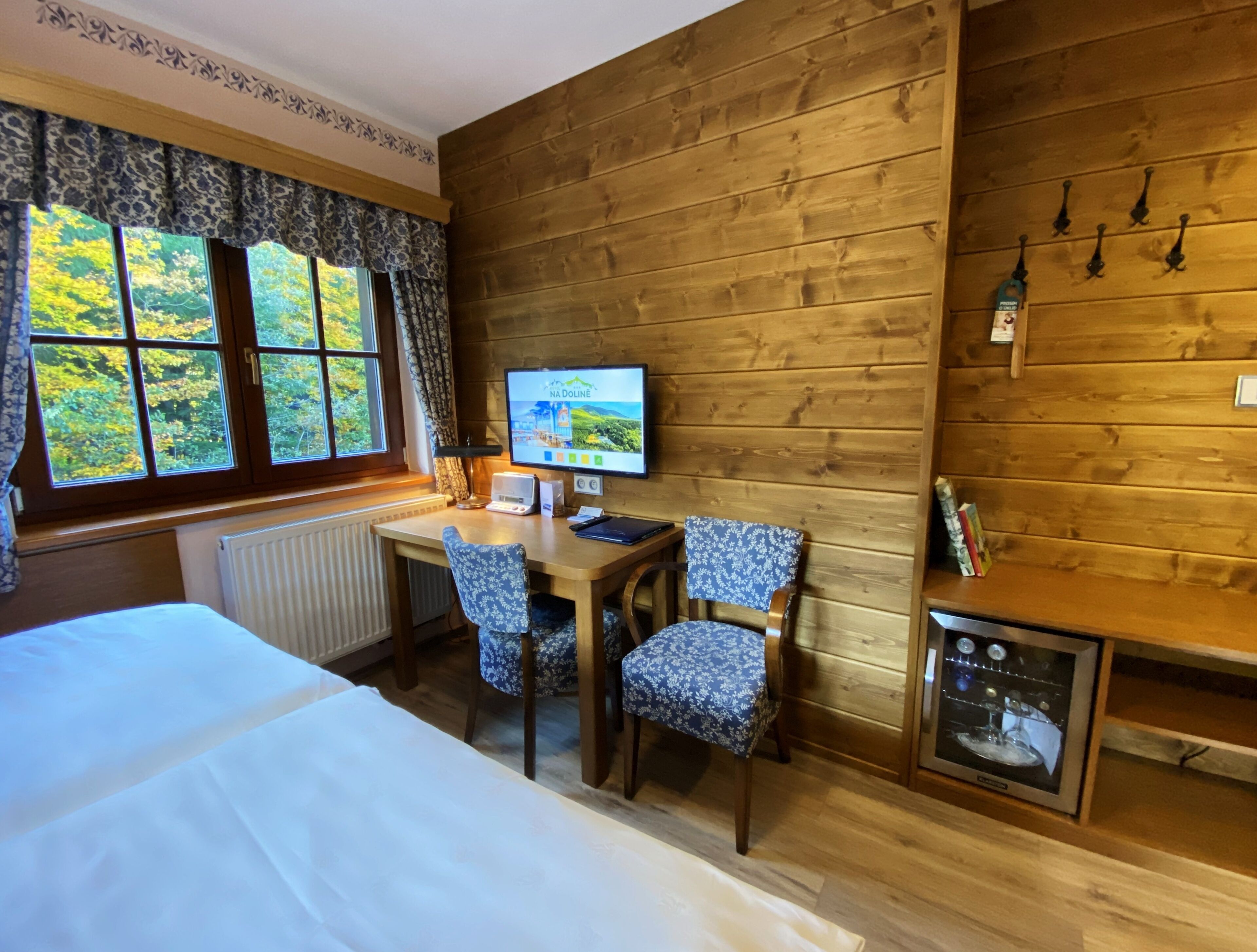 Ein gemütliches Zimmer mit Holzwand, einem Bett, einem Schreibtisch mit Computer, zwei gemusterten Stühlen und einem kleinen Fernseher.