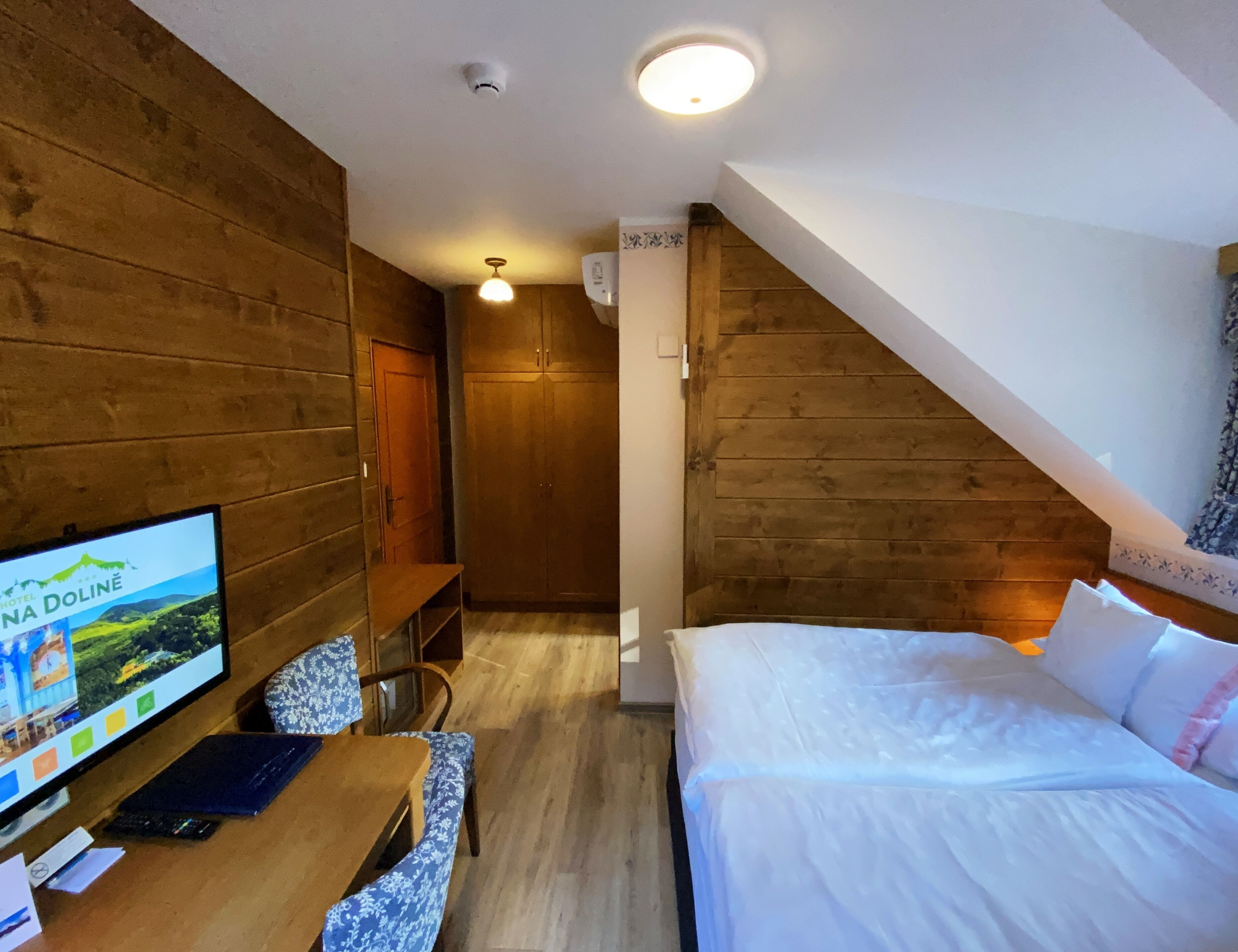Een hotelkamer met houten wanden, een bed, een bureau met een computer en een televisie waarop een landschap wordt getoond.