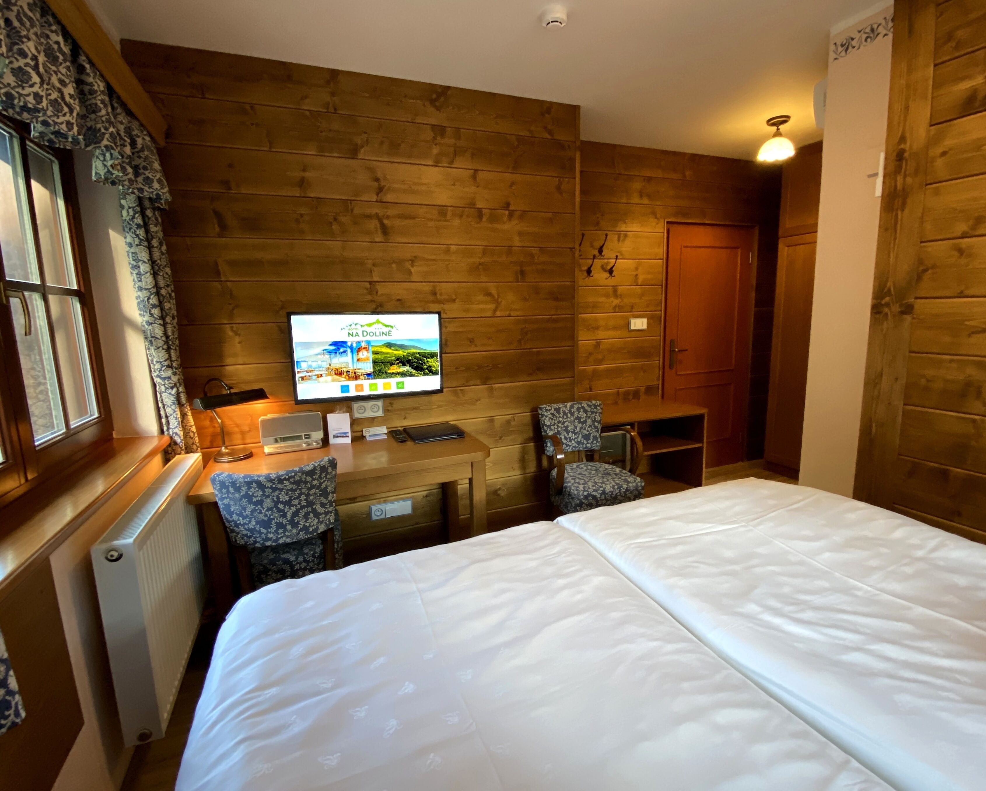 Habitación de hotel con pared de madera, cama, escritorio con silla, televisor y ventana con cortinas.