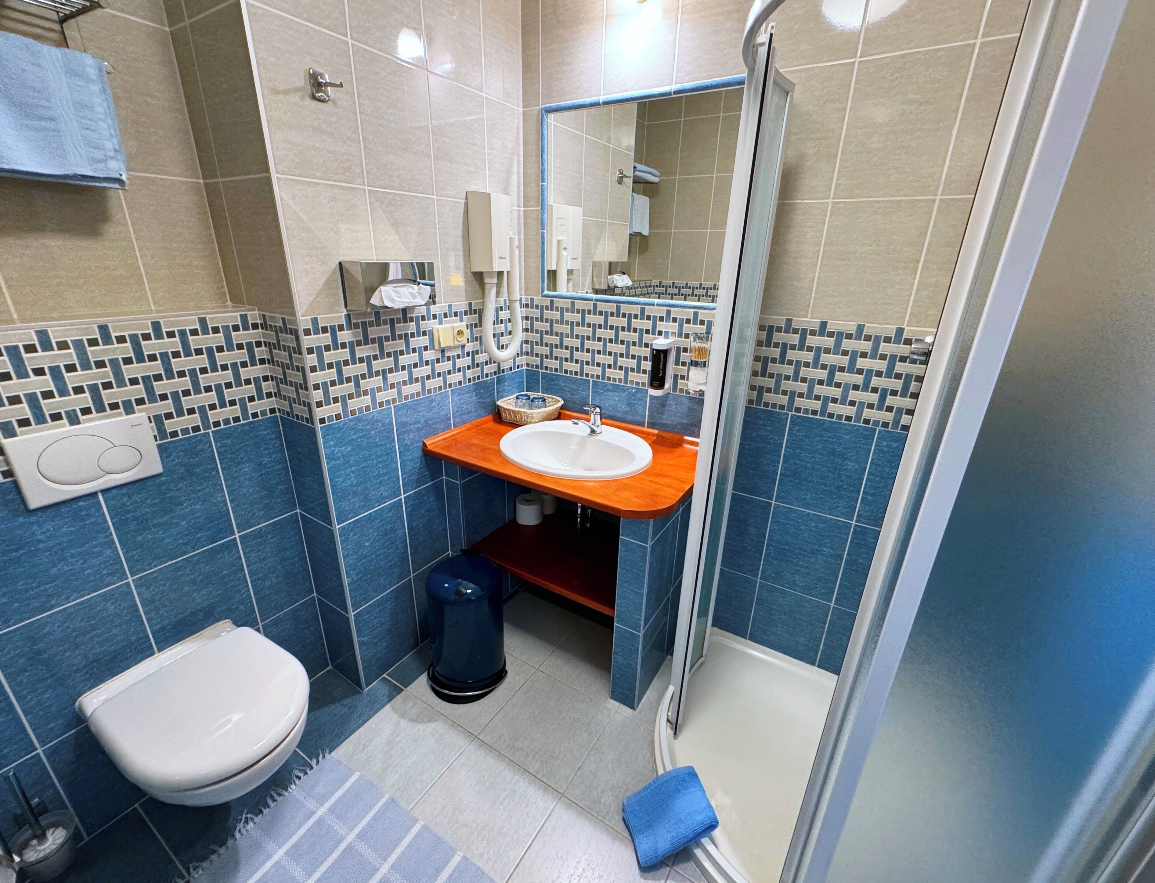 Un baño con inodoro blanco, lavabo con encimera azul, zona de ducha y suelo de baldosas.