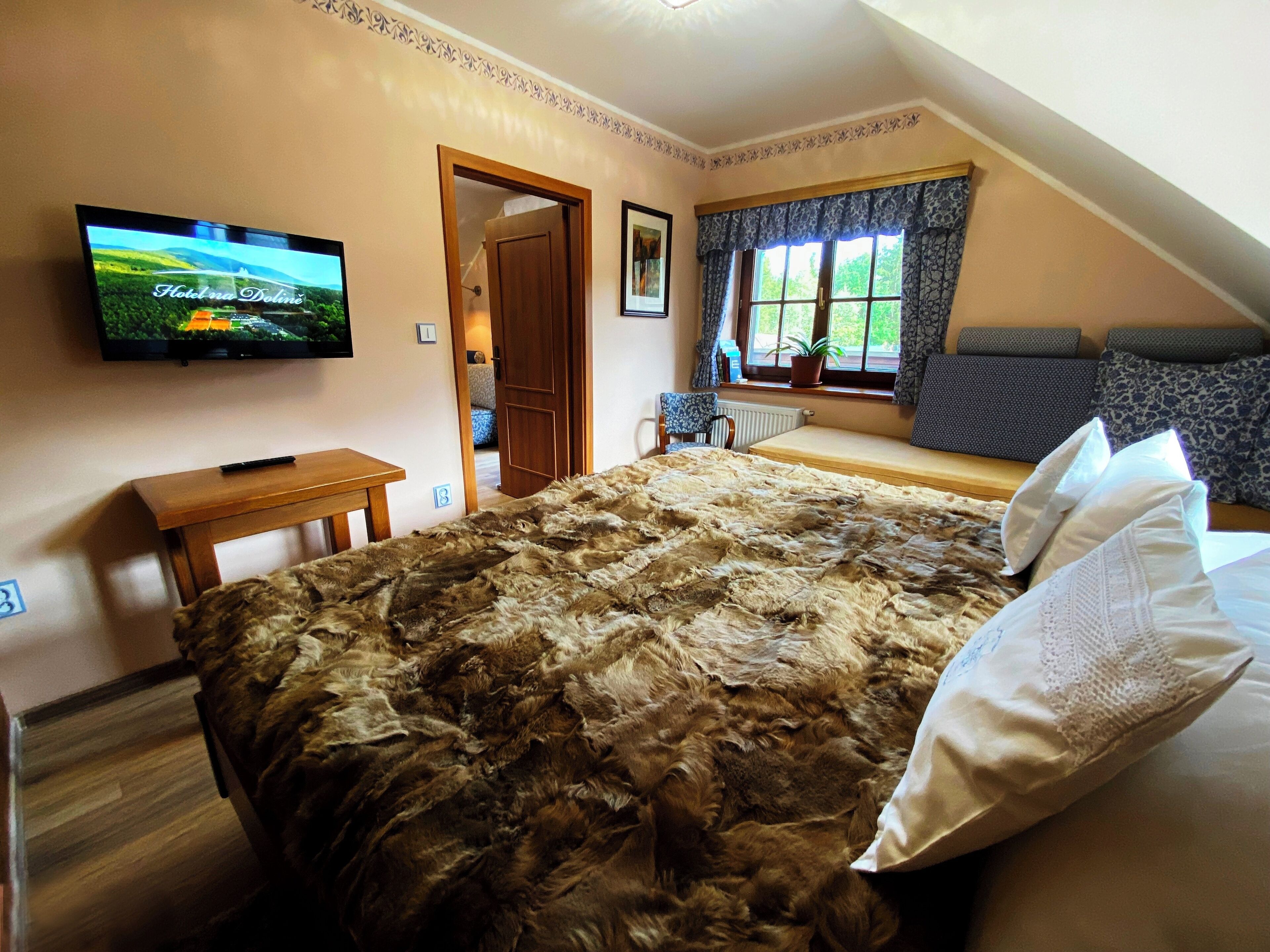 A bedroom with a large bed, a television mounted on the wall, a window with curtains, and a small table.