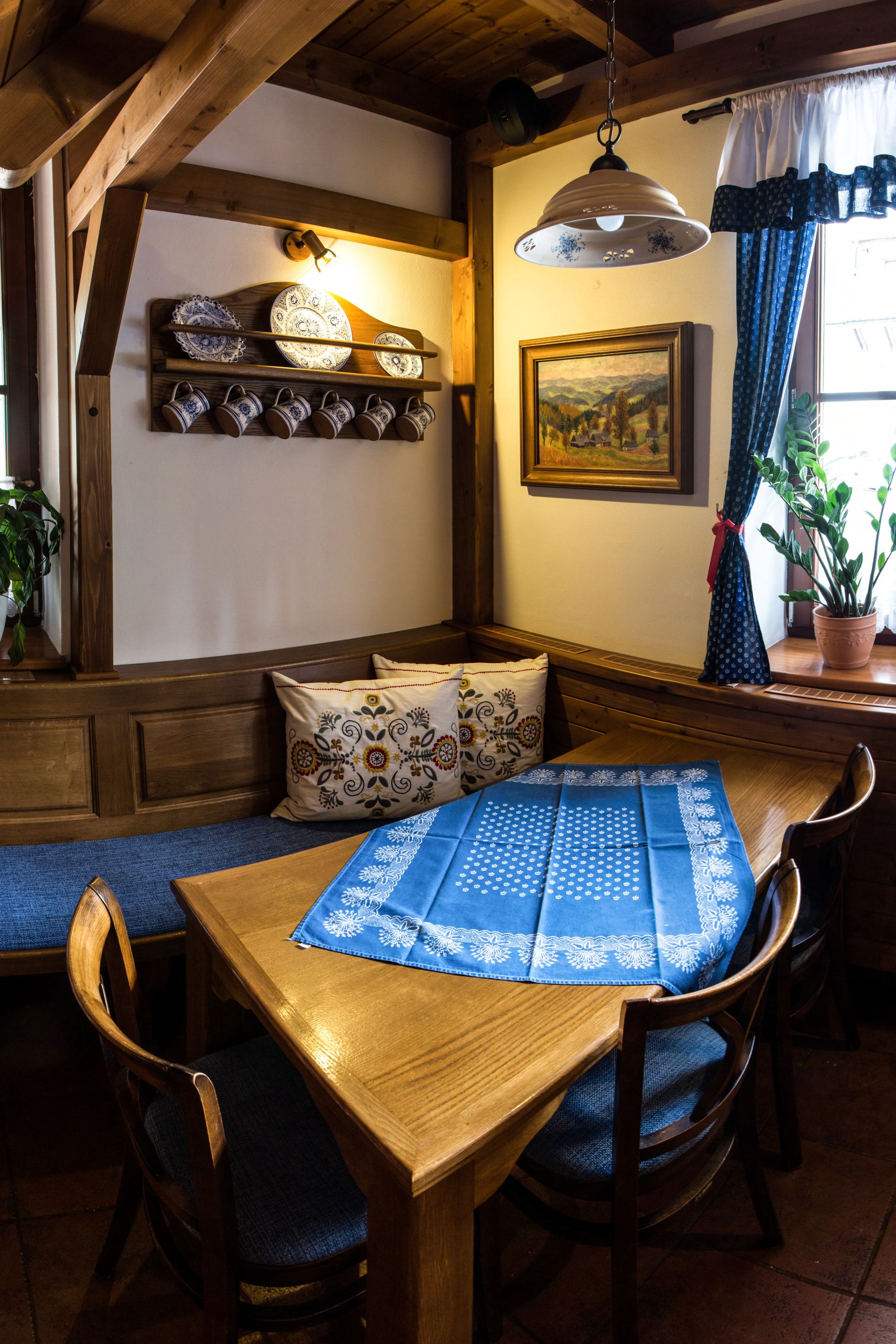A wooden dining table with chairs, a bench, and a blue tablecloth. There is a wall-mounted shelf with plates and cups, a hanging light fixture, and a framed painting.