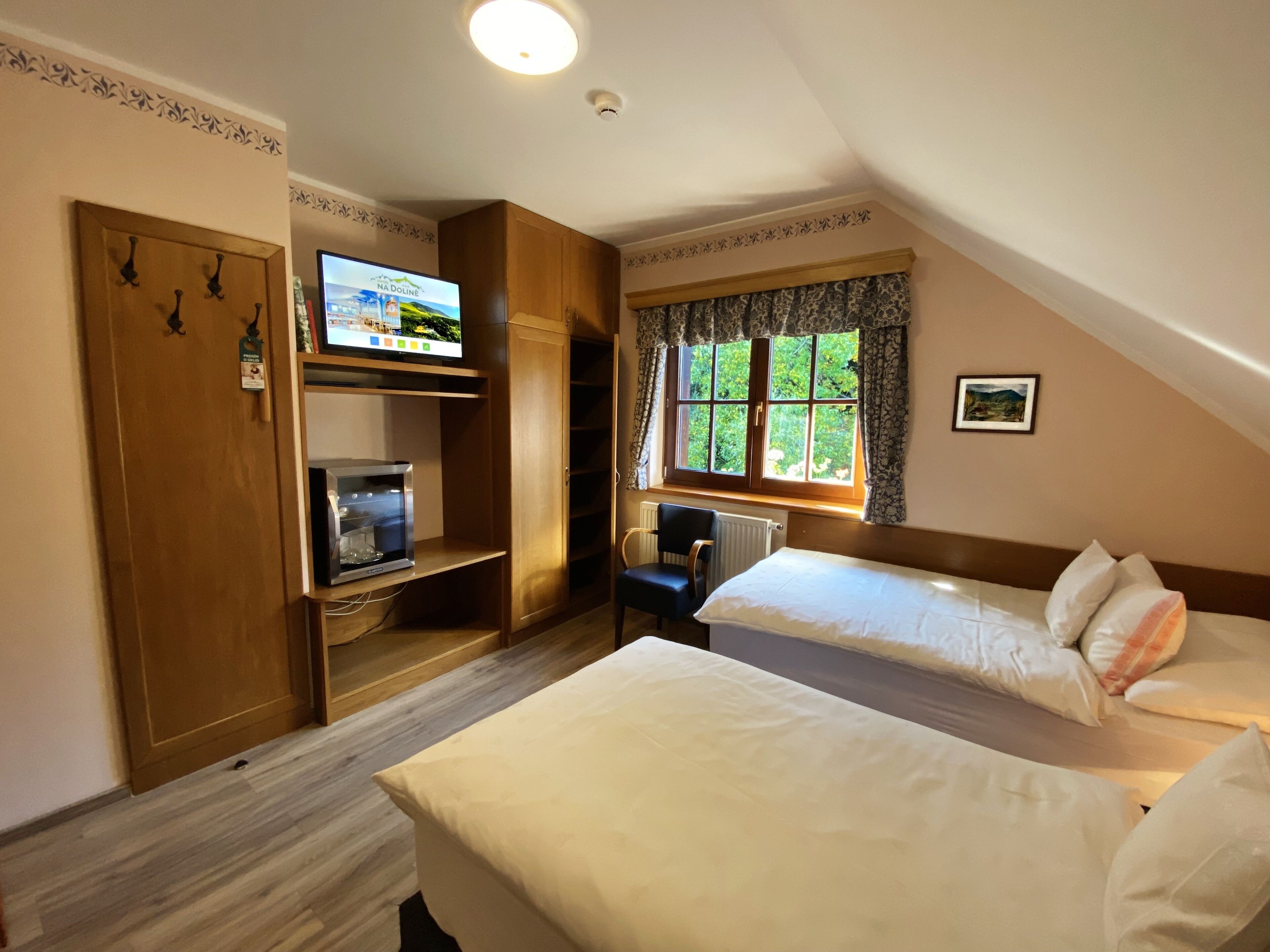 A bedroom with two beds, a television, a wooden wardrobe, and a window with a view of greenery.