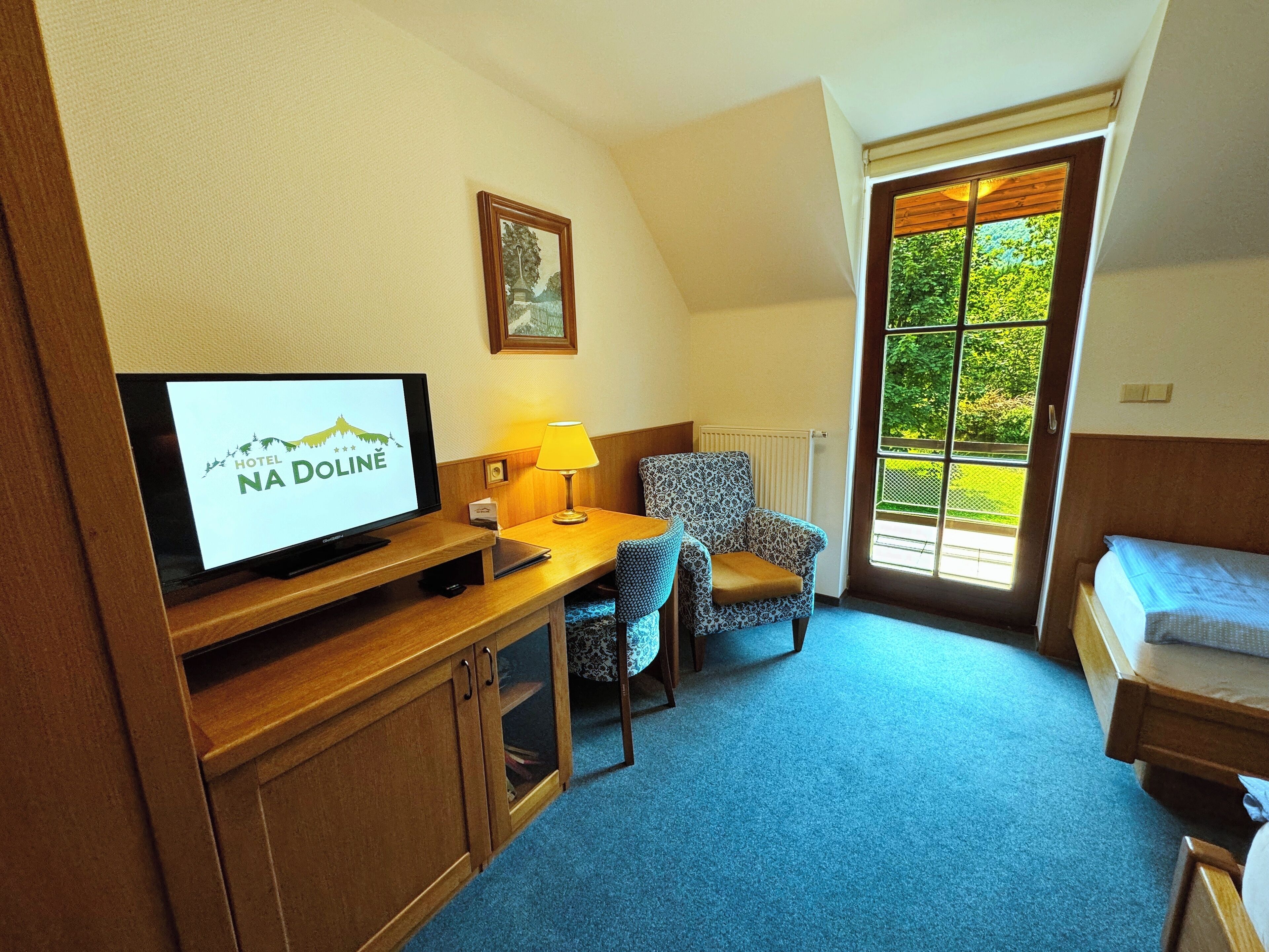 A hotel room with a television displaying the hotel's name, a desk with a lamp, a patterned chair, a bed, and a window with a view of greenery.