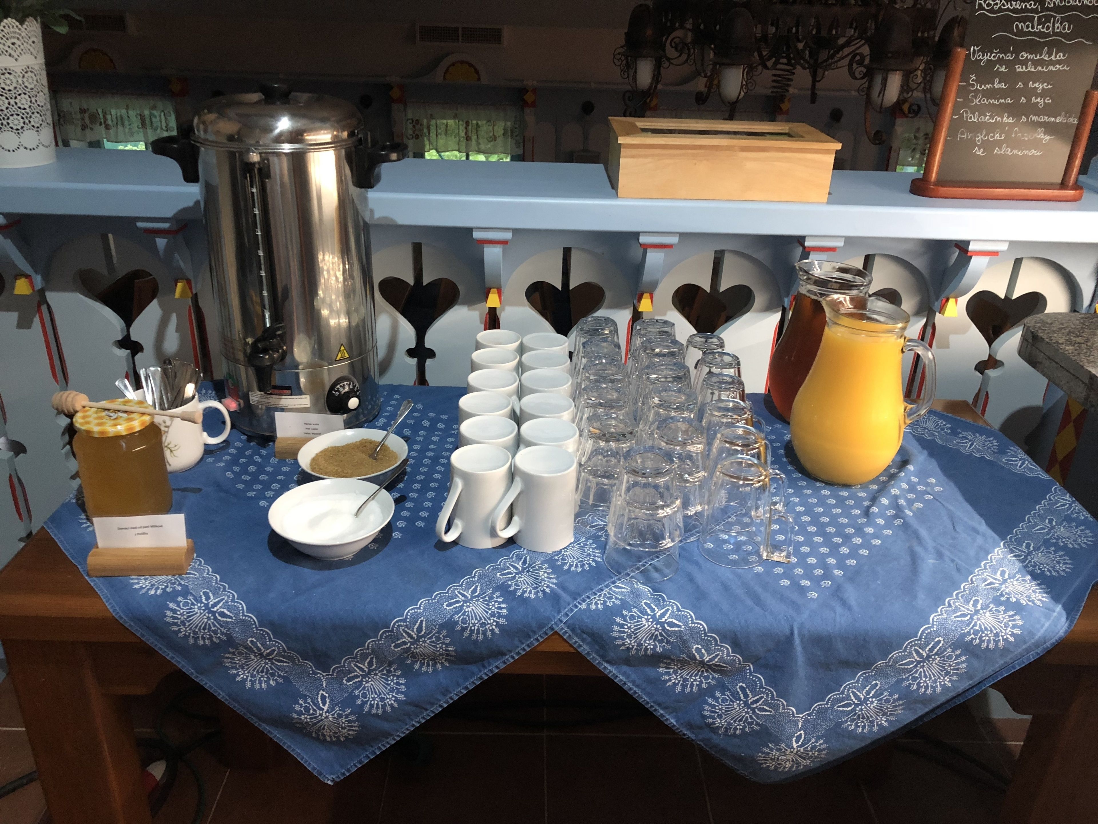 A table with a blue tablecloth, a coffee pot, sugar bowls, white mugs, and a pitcher of orange juice.