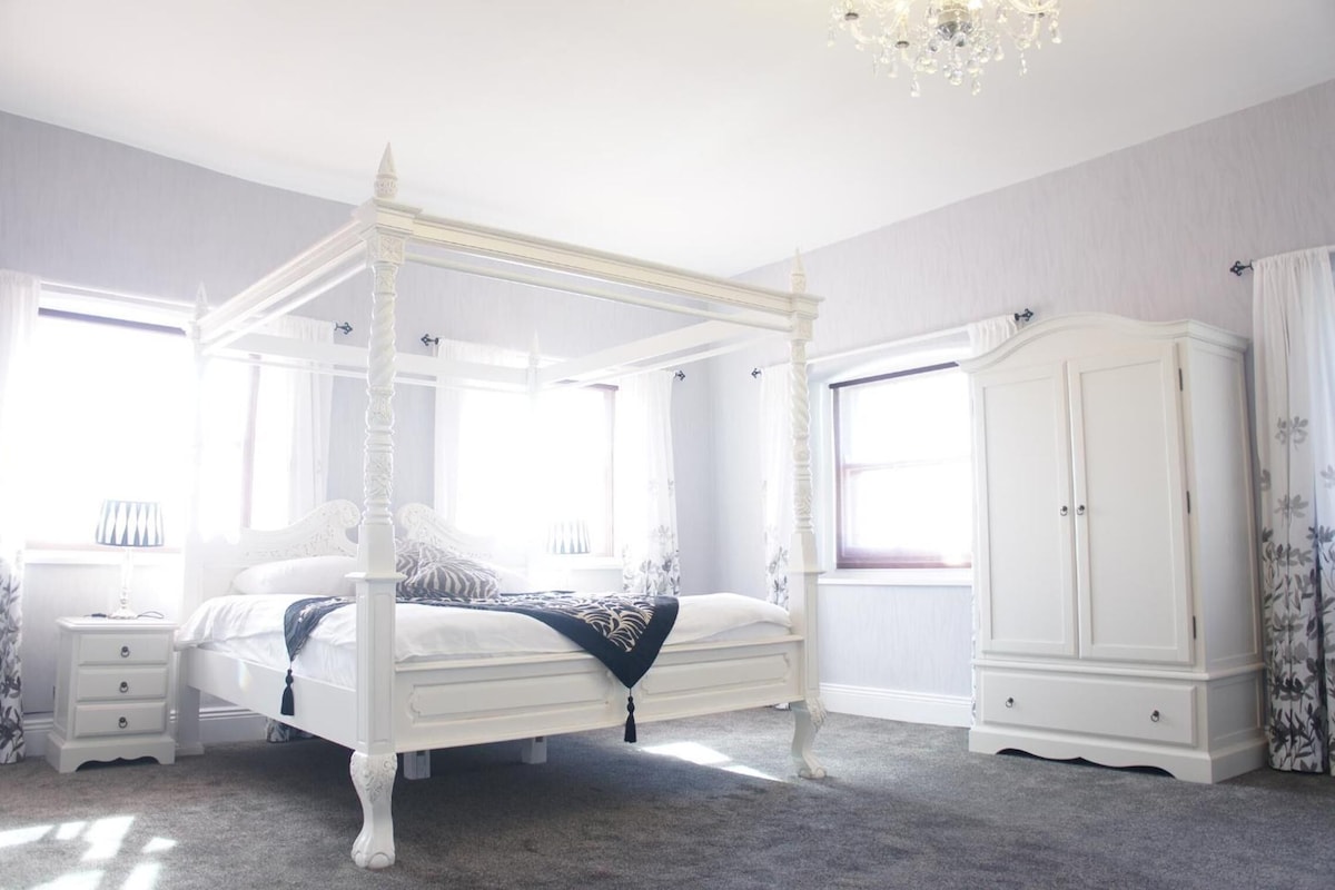 A four-poster bed with a canopy, a nightstand, a wardrobe, and a chandelier in a room with large windows.