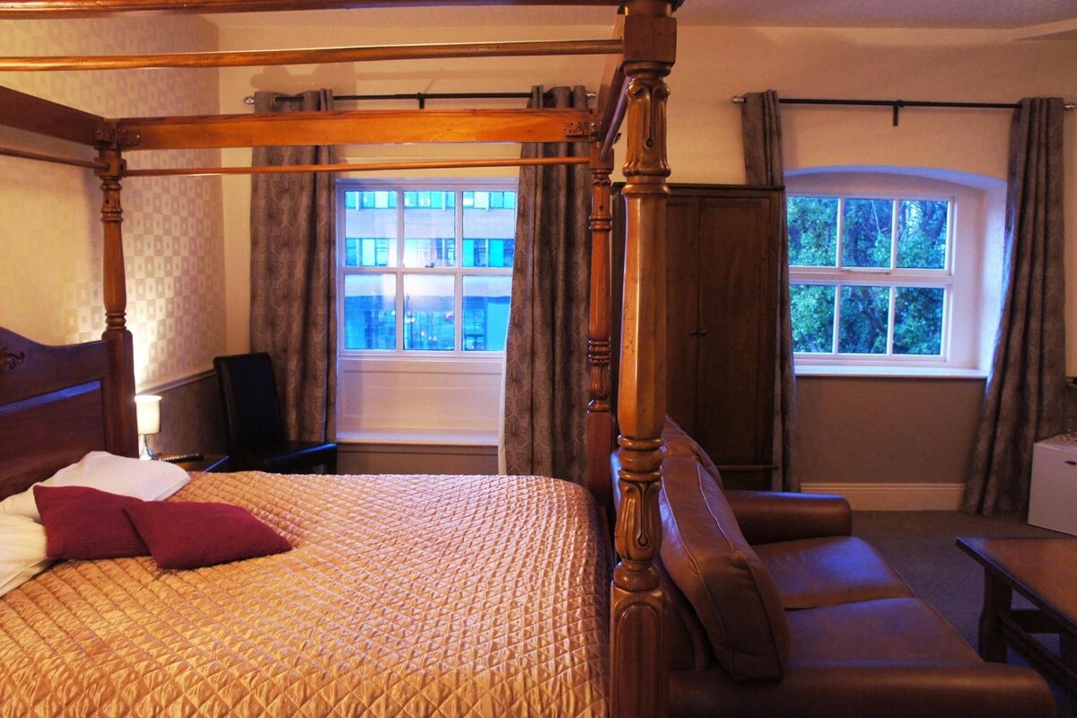 A four-poster bed with a quilted bedspread, a wooden headboard, and a nightstand with a lamp. There is a window with curtains and a view of buildings outside.