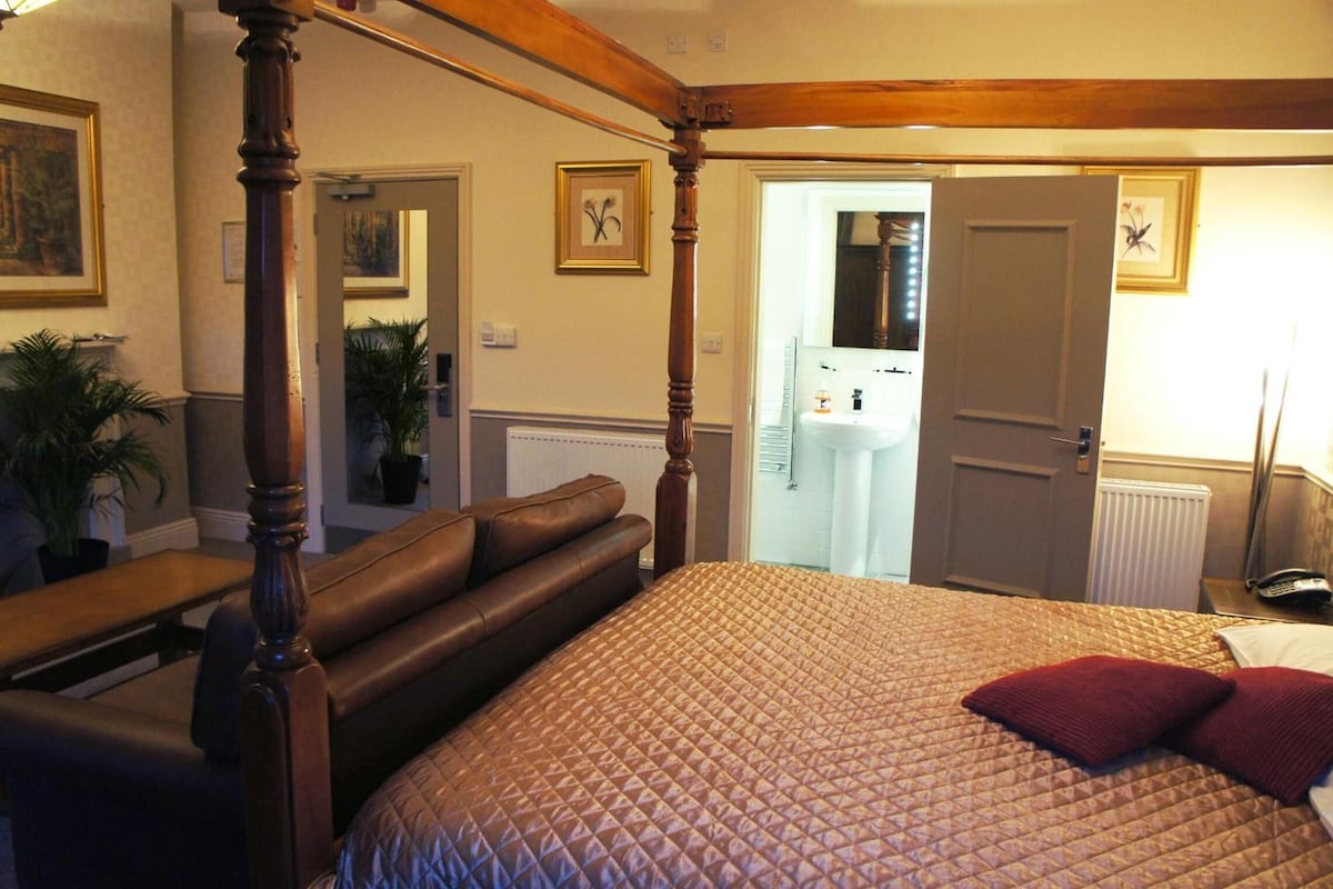 A four-poster bed with a quilted headboard, a bench, and a red pillow. There is a door leading to a bathroom with a mirror and a sink.