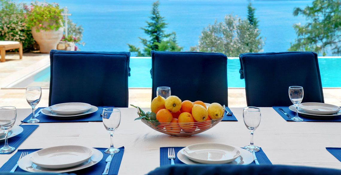A table set for three with a bowl of fruit in the center, overlooking a pool and the sea.
