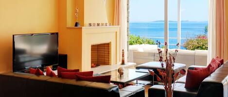 Un salon avec un téléviseur, un canapé et une cheminée. De grandes fenêtres offrent une vue sur la mer.
