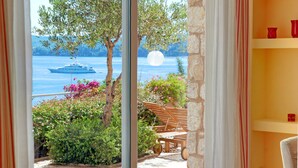 A room with a view of a lake, a boat, and a moon. There are plants and a wooden bench outside the window. - Corfu Luxury Villas (Corfu)