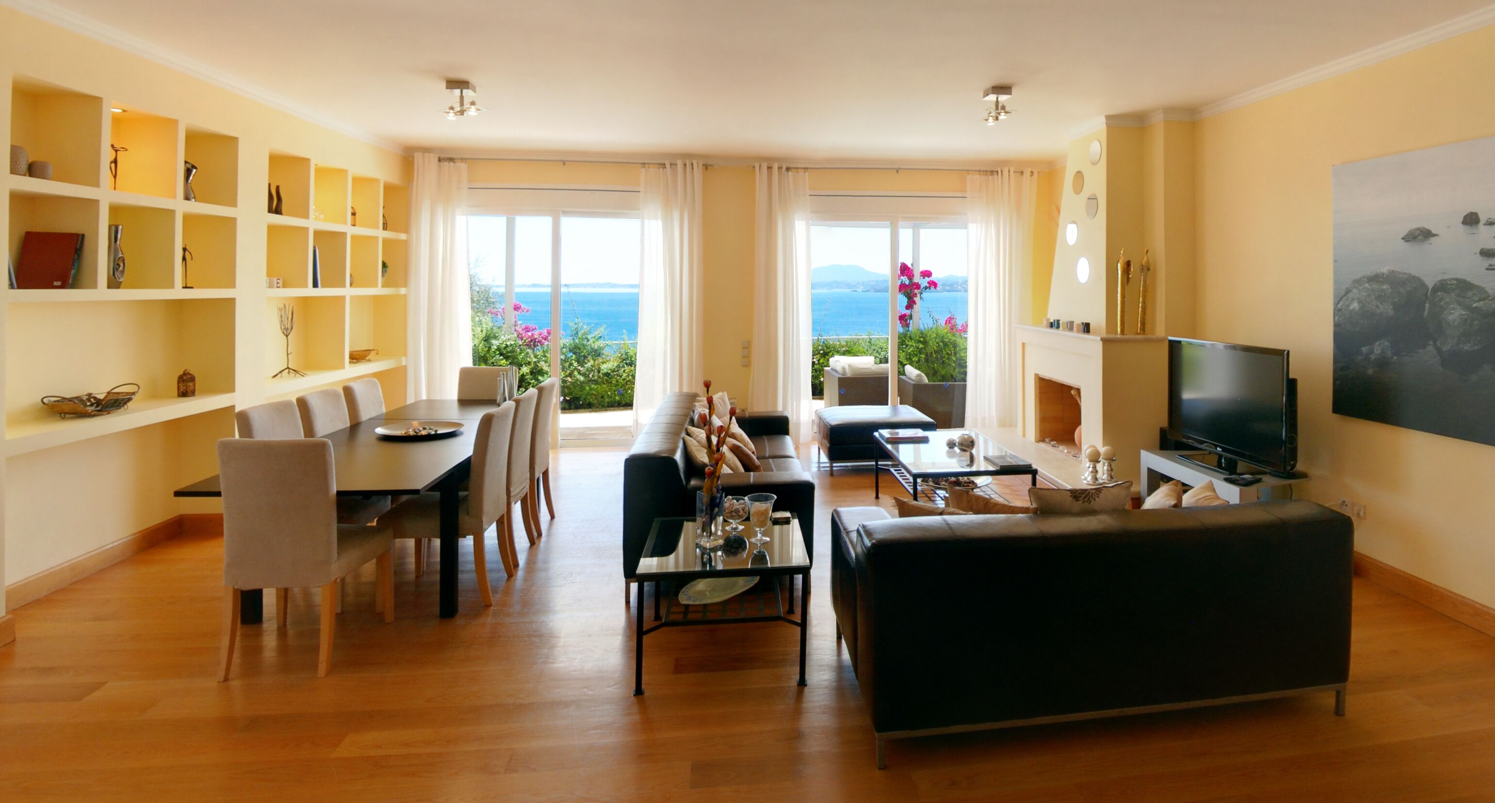 Amplia sala de estar con comedor, sofá, televisor y chimenea. Hay un ventanal con vista al mar.
