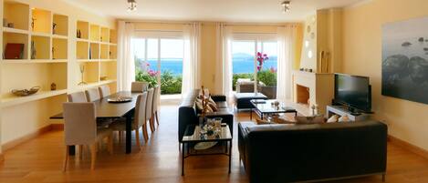 Amplia sala de estar con comedor, sofá, televisor y chimenea. Hay un ventanal con vista al mar.