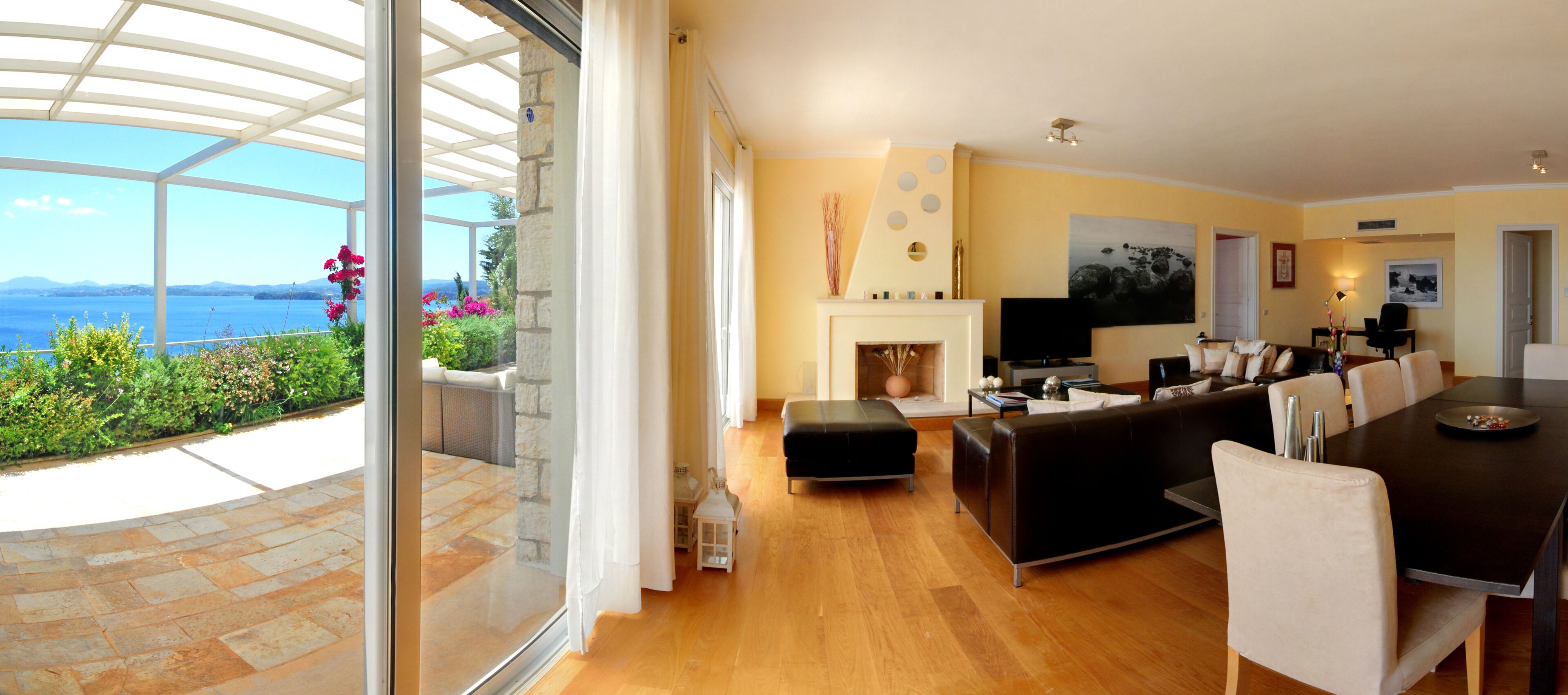 A modern living room with a fireplace, a dining area, and a large glass door leading to a patio with a view of the sea.