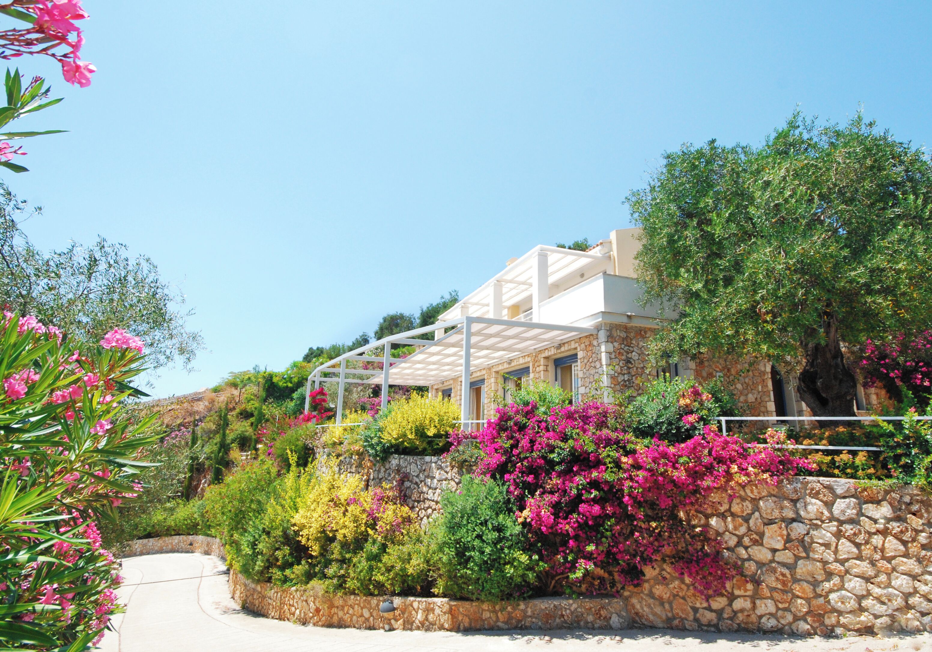 A stone building with a white balcony, surrounded by lush greenery and pink flowers.