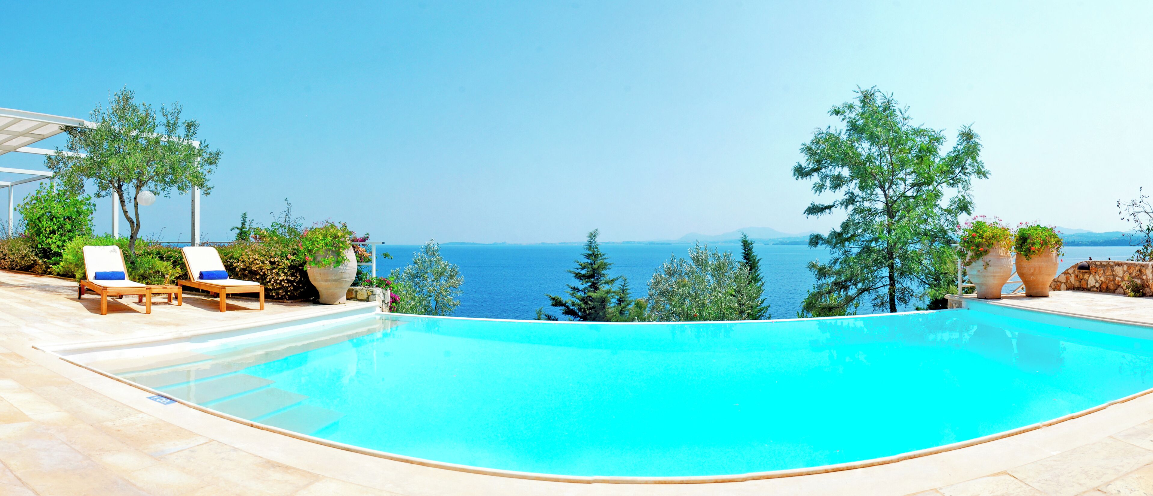 A pool with a clear view of the sea, surrounded by lounge chairs and potted plants.