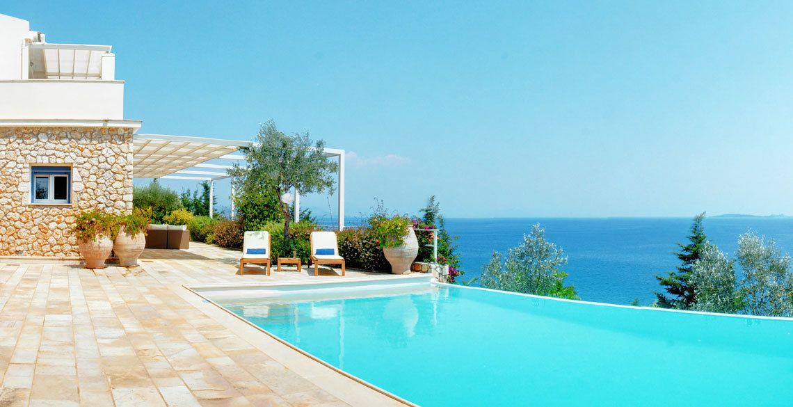 A modern building with a stone facade, a swimming pool, and outdoor seating with a view of the sea.