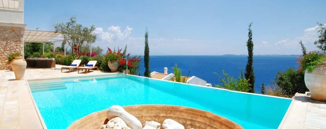 A pool with clear blue water, surrounded by lounge chairs and potted plants, overlooking a scenic view of the sea.