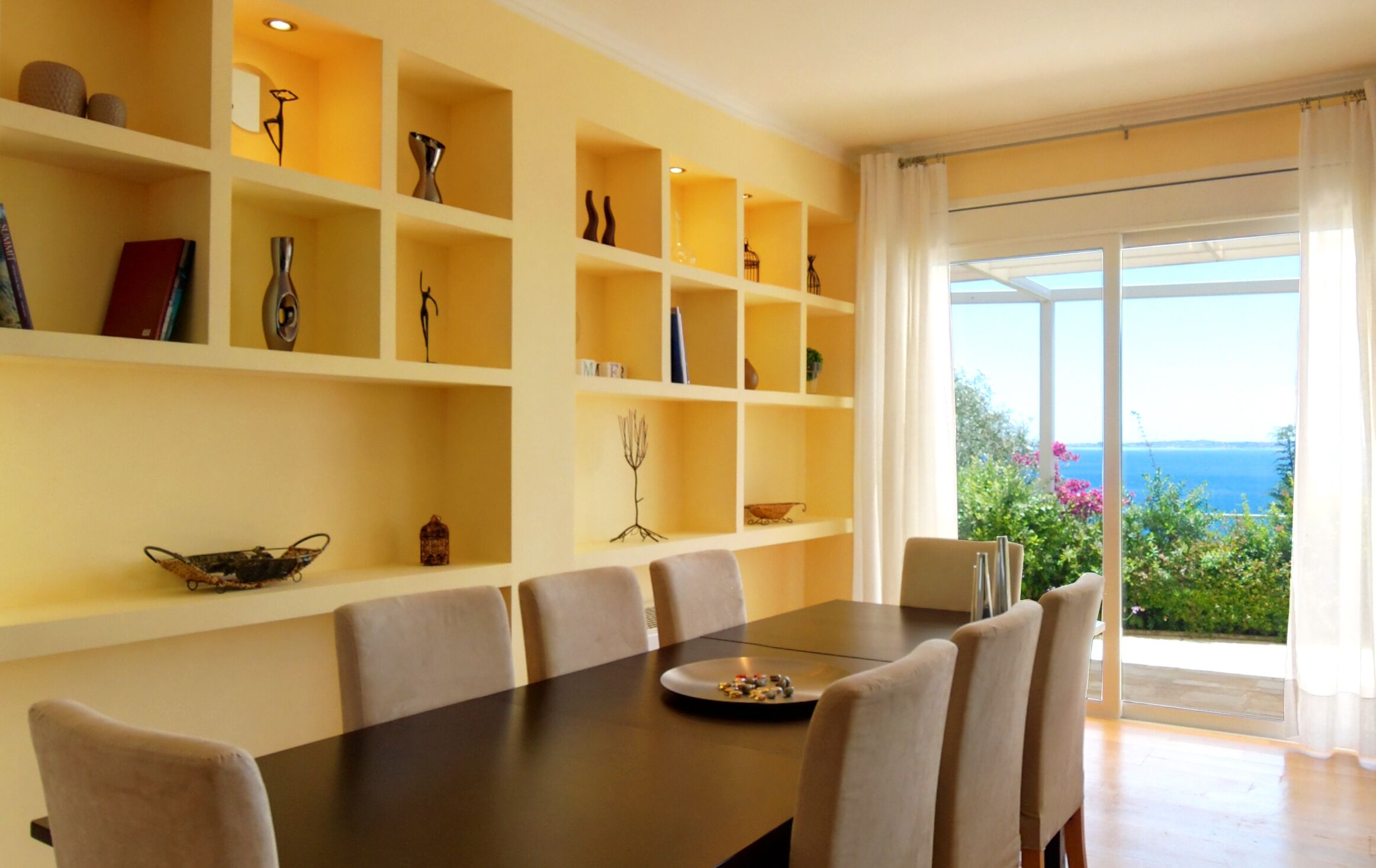 A dining area with a dark wooden table, beige chairs, and a view of the sea through large windows.