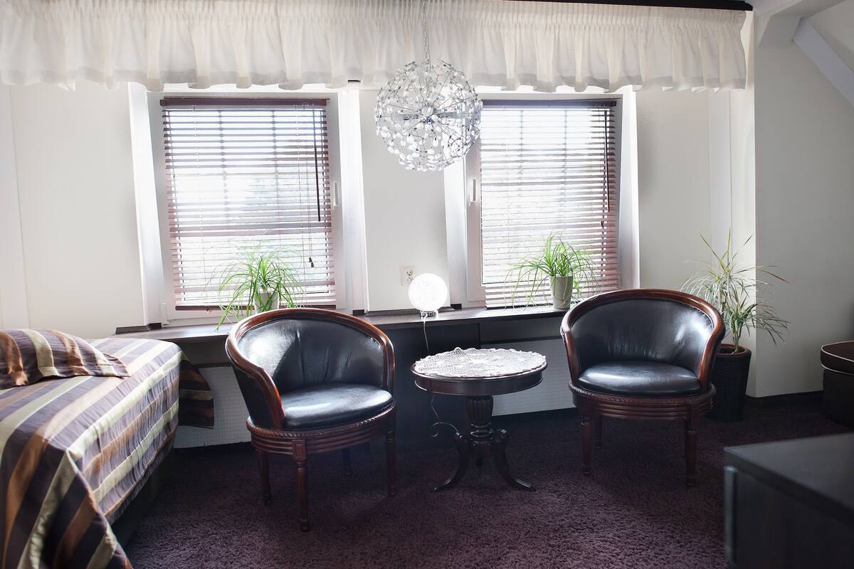 A bedroom with a bed, two leather chairs, a small table, and two windows with blinds.