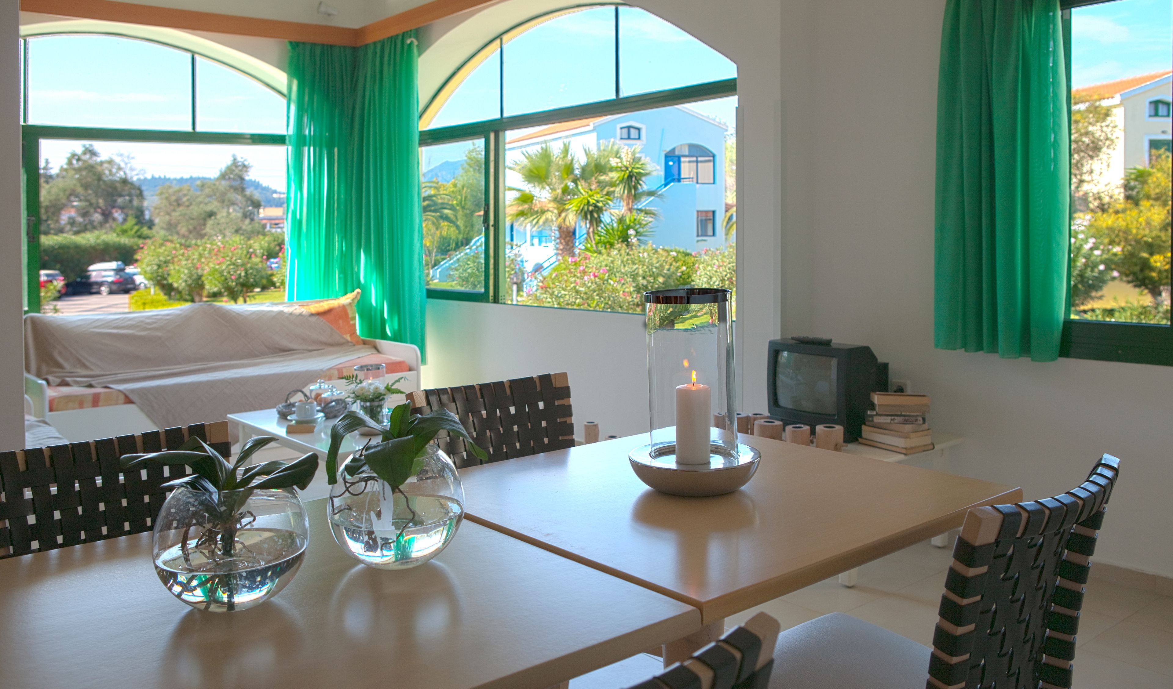 A room with a dining table, chairs, a TV, and a view of a blue house and greenery outside.